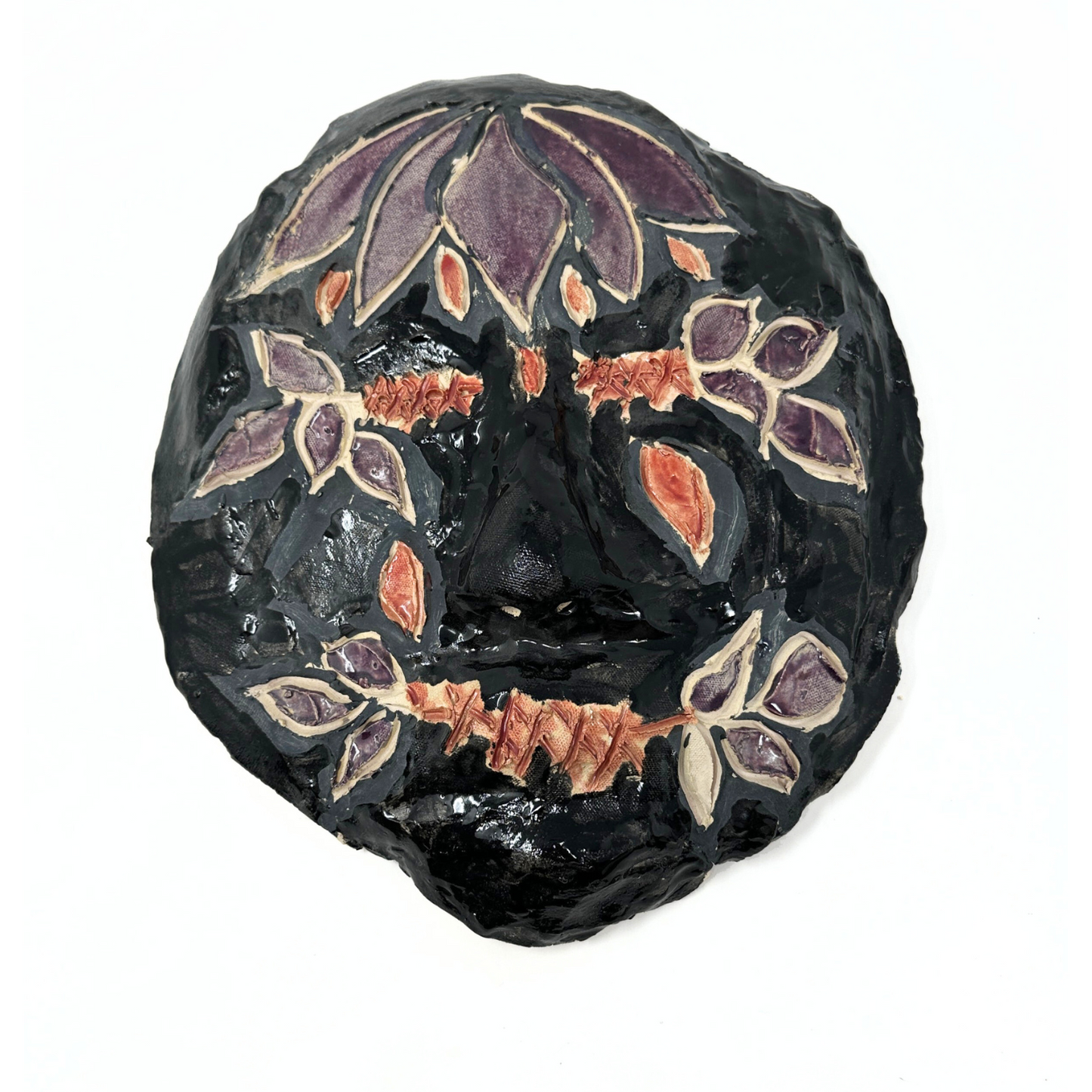 a ceramic mask sculpture in the shape of a round head. The surface of the face is glazed mostly in black, with purple flowers and leaf designs near the eyes, mouth, and across the forehead. The eyes and mouth of the face appear to be stitched shut, and the etched stitch marks are painted red. There are several red tear shaped petals on the cheeks. 