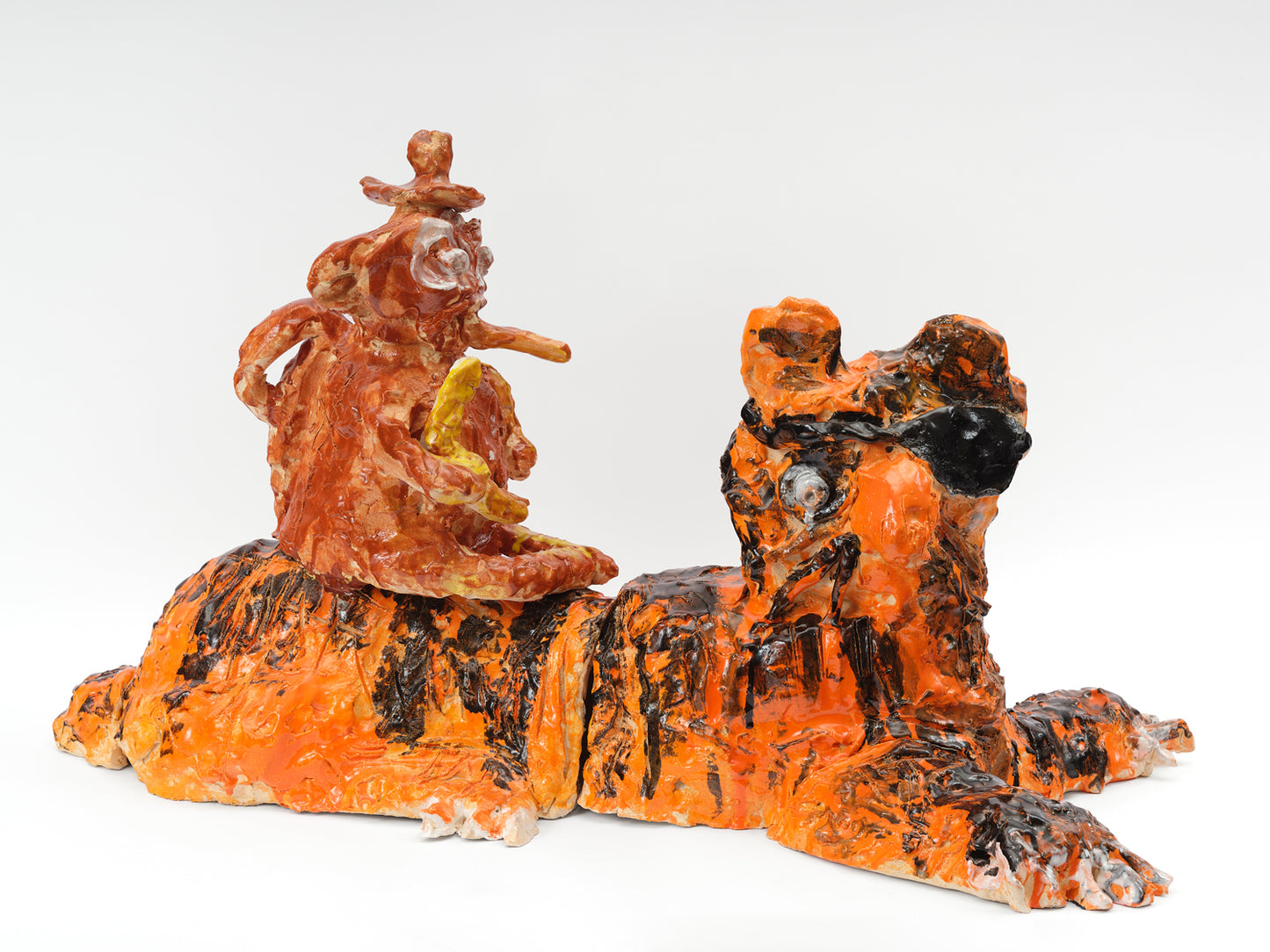 A large ceramic sculpture of a tiger with a monkey crouched on its back. The tiger is glazed bright orange with black stripes. It's sitting down and wearing an eye patch. The monkey is glazed all light brown. It holds a banana, smokes a cigar, and wears a hat. 