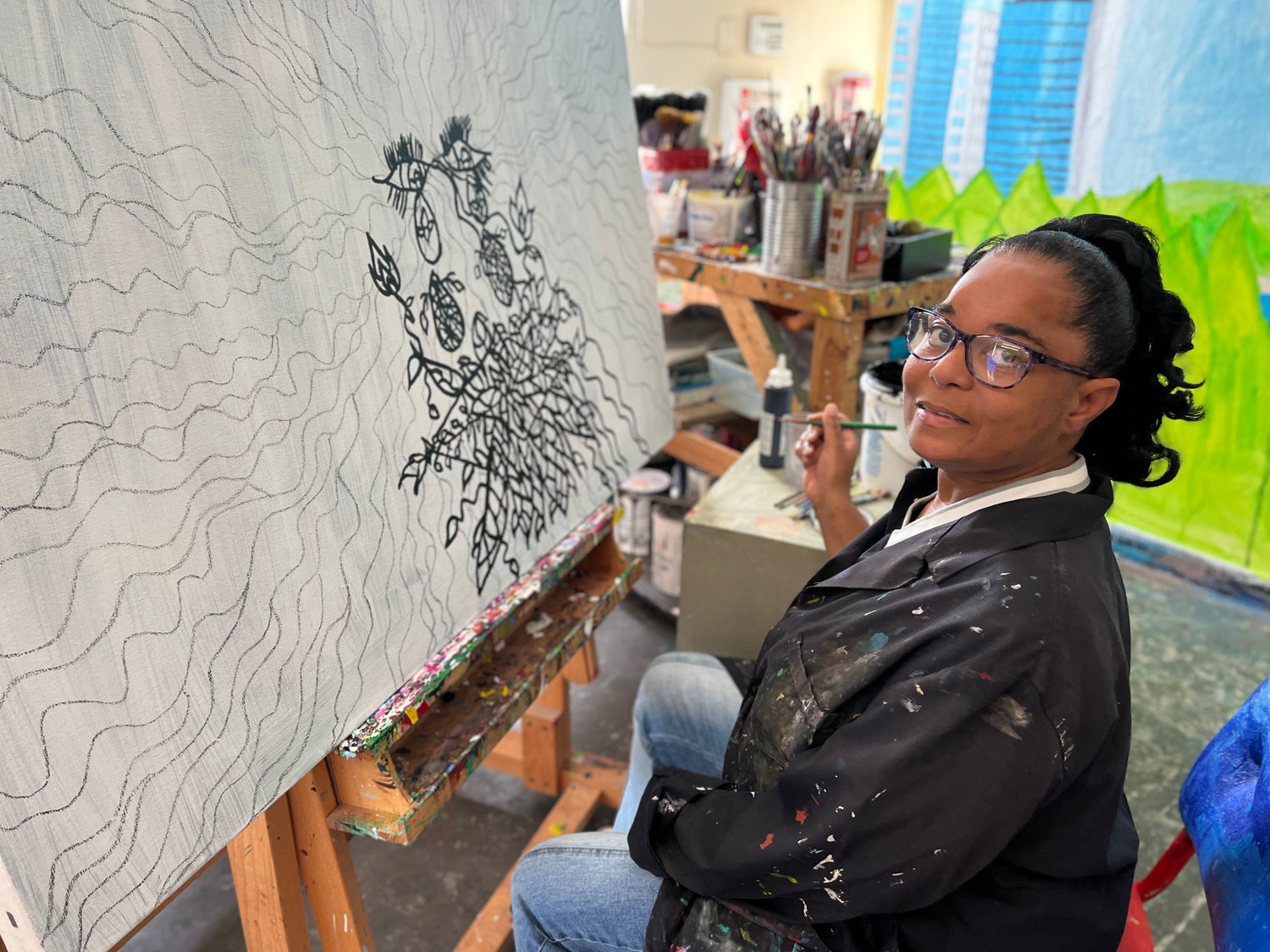 A photograph of an artist sitting in a painting studio. The artist is a black woman with glasses and long dark hair pulled into a pony tail. She looks over her shoulder and smiles at the camera. She wears a black shirt splattered with paint, and holds a small paintbrush in her right hand. A large white canvas is propped on an easel before her. The canvas is filled with wiggling lines and floral designs, most drawn in pencil and some traced over with black paint.