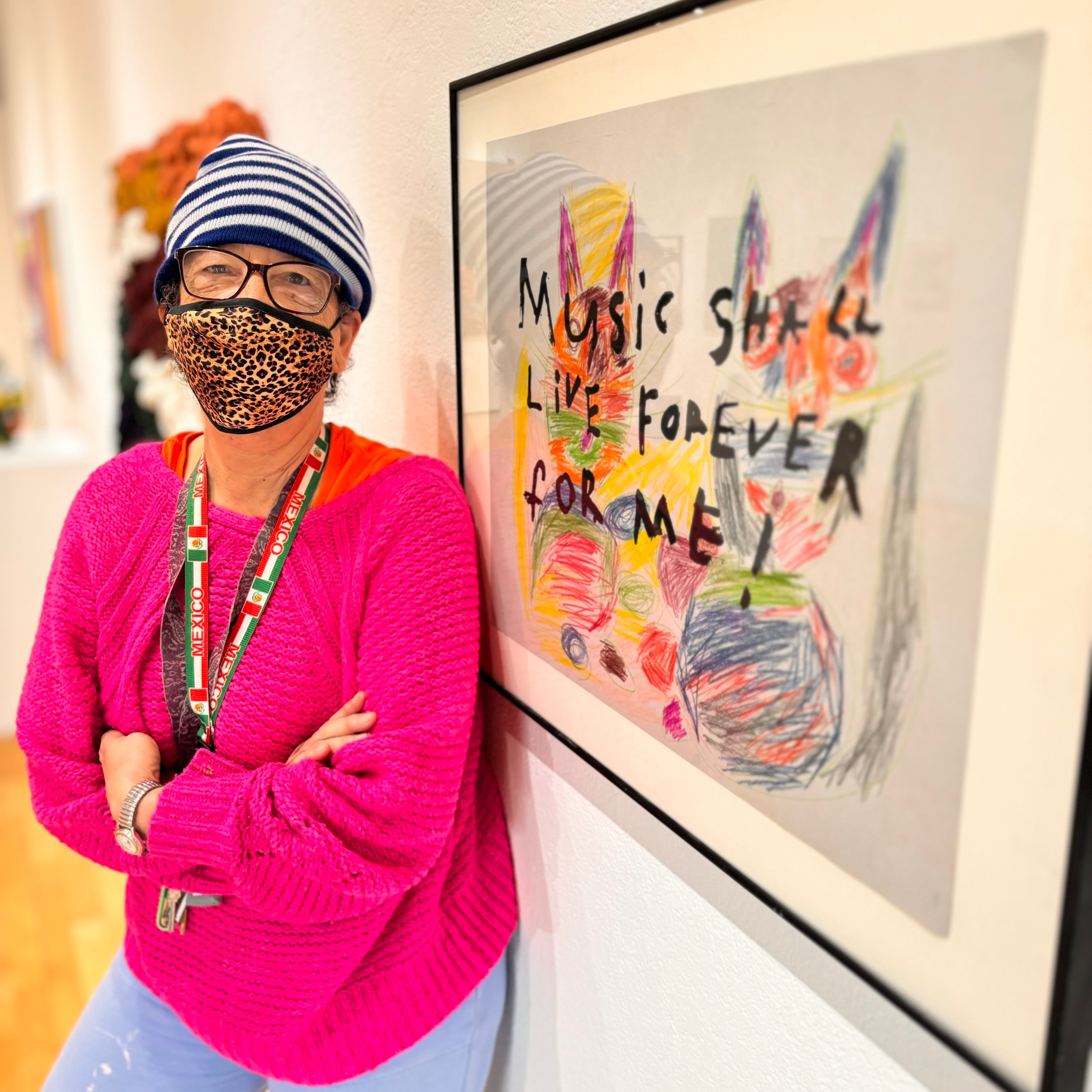 Dorian Reid, an artist with light brown skin, wears a bright pink sweater, a leopard print mask, and a striped beanie. She's leaning against a wall with her framed artwork that says "Music Shall Live Forever For Me!"