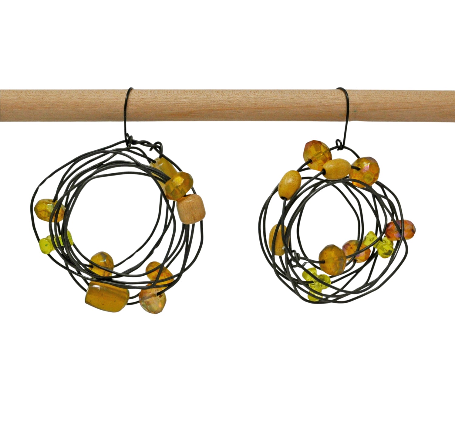 A set of unique handmade earrings hanging from a wooden dowel. The earrings are made from coiled black wire and beads. The design of each earring is concentric wire circles that have been sparsely strung with brown, amber, and yellow beads. Some of the beads are wooden and some are shiny translucent plastic.