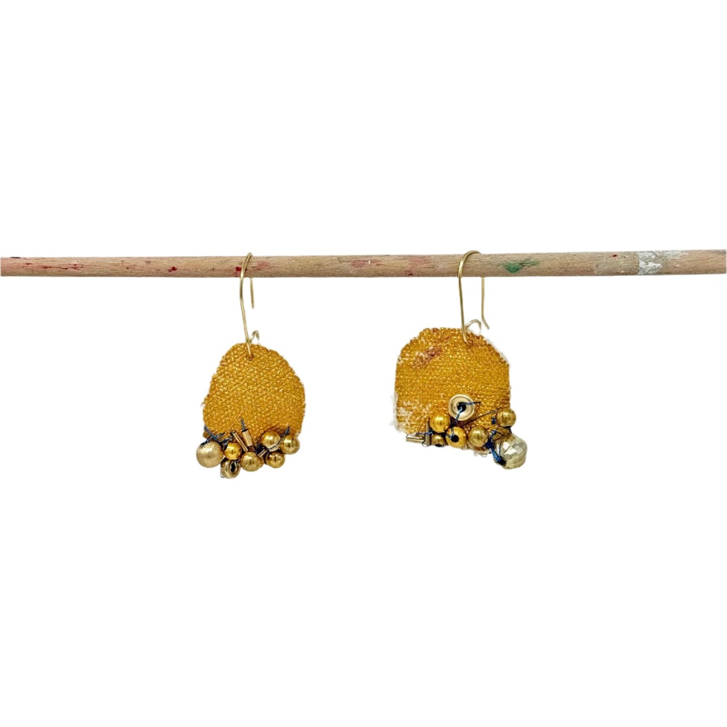 A pair of handmade earrings hanging form a wooden dowel, photographed in a white space. The earrings are round pieces of canvas, painted gold, with clusters of metallic beads stitched along each bottom edge.