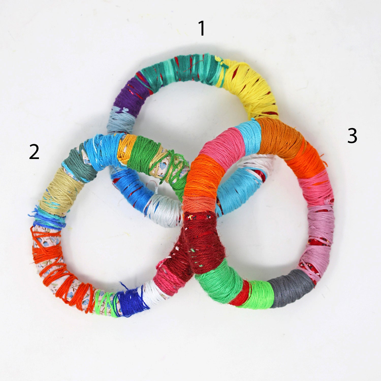 Another image of the bracelets, with numbers written by each one for shopping purposes. The bracelet with the number 1 features stripes in mostly purple, blue, white, turqoise, and yellow. The bracelet labeled with number 2 is mostly orange, green, grey, white, and red. The bracelet labeled number 3 is mostly pink, grey, green, and orange. 