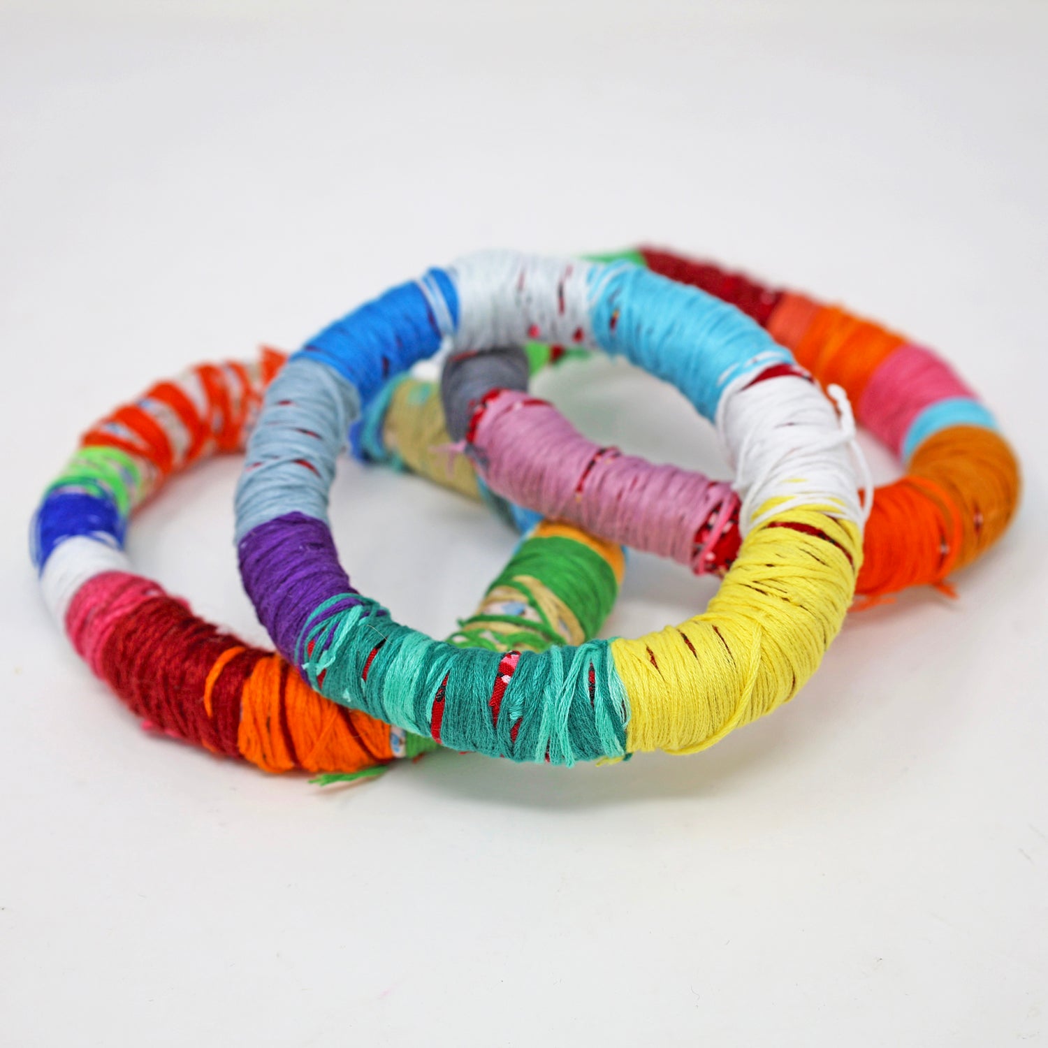 Three round bangle bracelets wrapped in embroidery thread and fabric. Each is striped with several bars of different colors: blue, purple, green, yellow, red, or orange. 