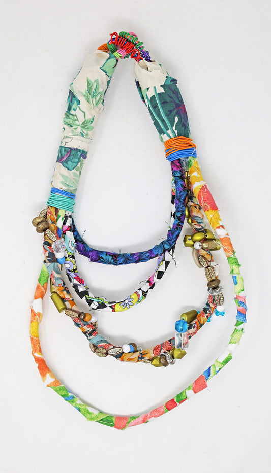 A mixed media statement necklace made from cords that have been wrapped with strips of fabric, wire, and beads. The necklace has four tiers, each bound with a different summery, floral patterned fabric. One of the tiers has a string of large plastic beads stitched around it in a spiral. 