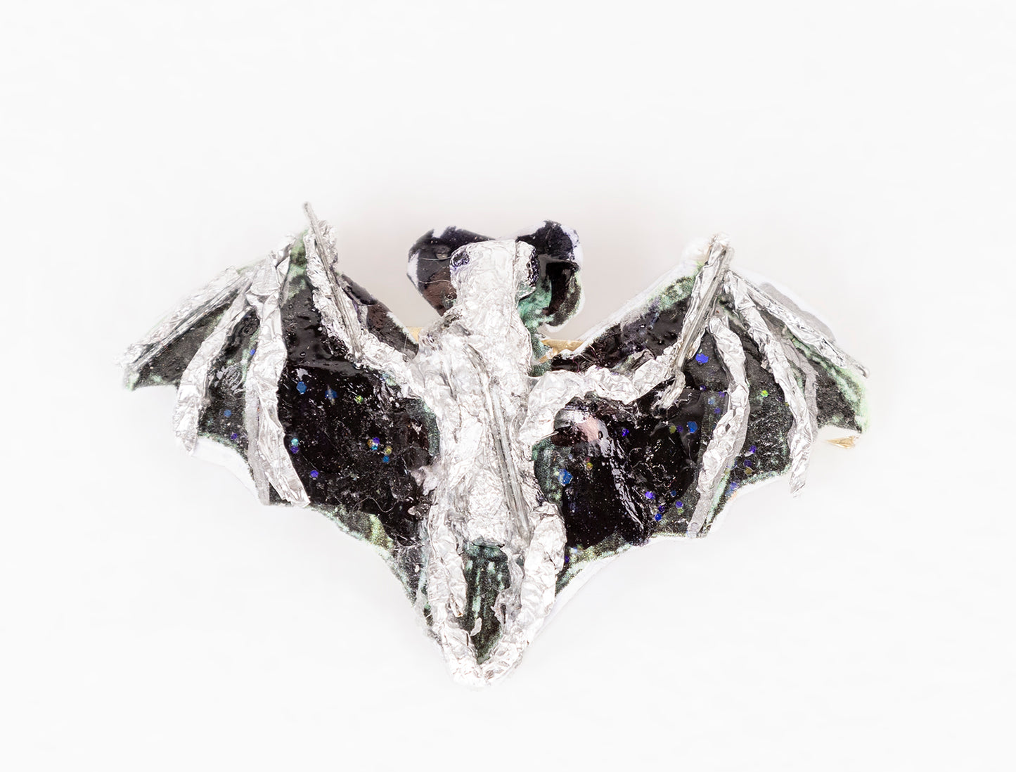a brooch shaped like an animal: a bat in flight. The bat is made with various media including wire, tape, glitter, paint, and other recyclable materials. The body of the bat is painted silver, and the wings are painted black with blue and green glitter sprinkled over the surface.  Photographed from this angle, the bat seen from above, facing away from the camera.