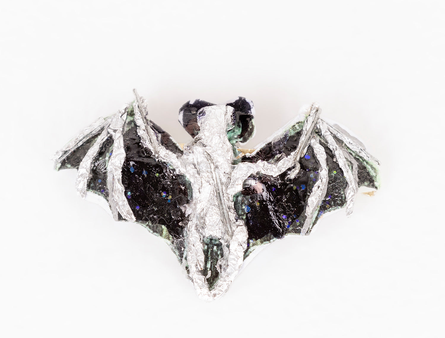 a brooch shaped like an animal: a bat in flight. The bat is made with various media including wire, tape, glitter, paint, and other recyclable materials. The body of the bat is painted silver, and the wings are painted black with blue and green glitter sprinkled over the surface.  Photographed from this angle, the bat seen from above, facing away from the camera.