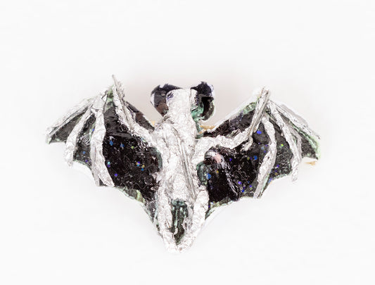 a brooch shaped like an animal: a bat in flight. The bat is made with various media including wire, tape, glitter, paint, and other recyclable materials. The body of the bat is painted silver, and the wings are painted black with blue and green glitter sprinkled over the surface.  Photographed from this angle, the bat seen from above, facing away from the camera.
