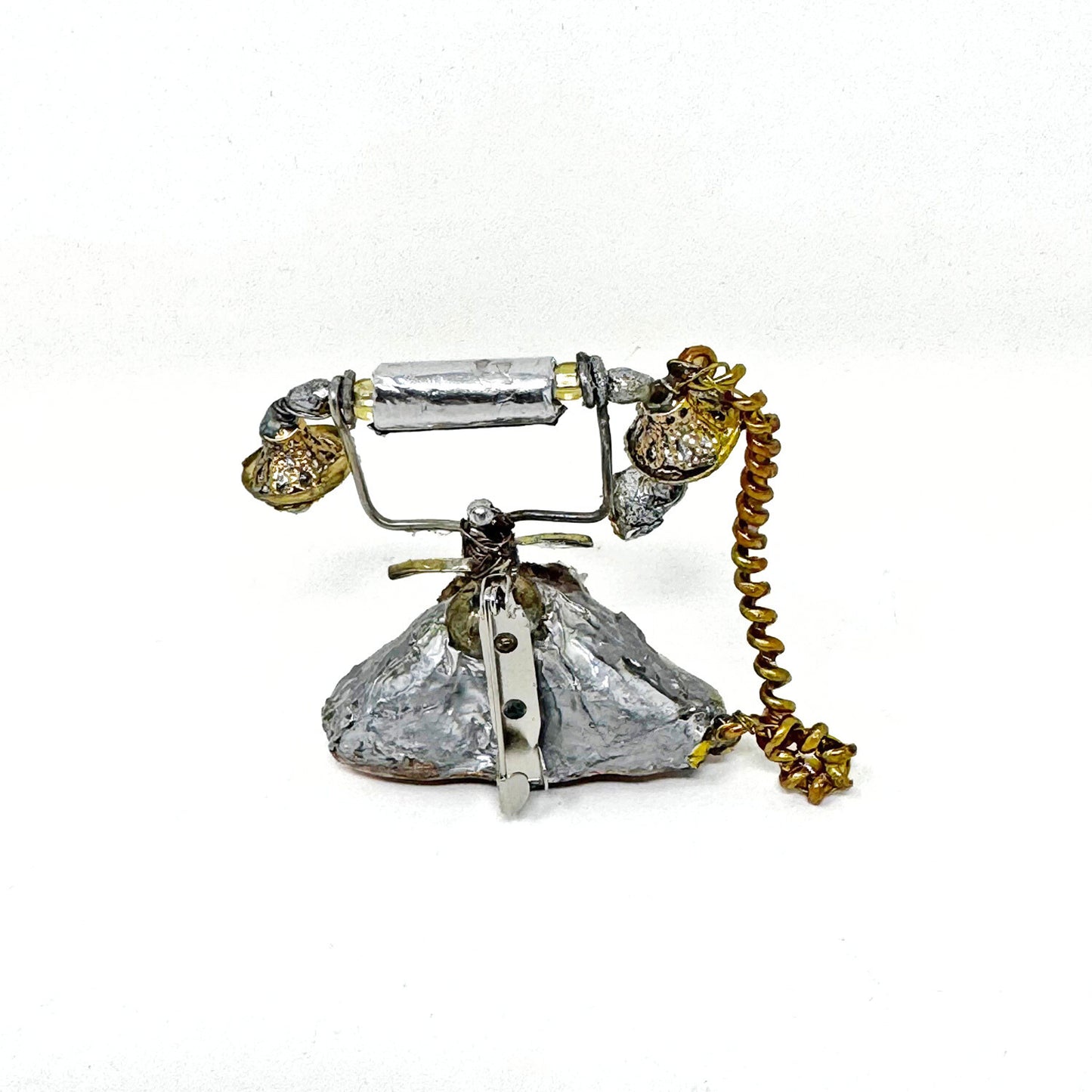 Backside of a mini sculpture of a silver antique style rotary phone, made of recycled materials against a white background. There is a metal pinback attached to the sculpture  that allows it to be worn as a brooch.