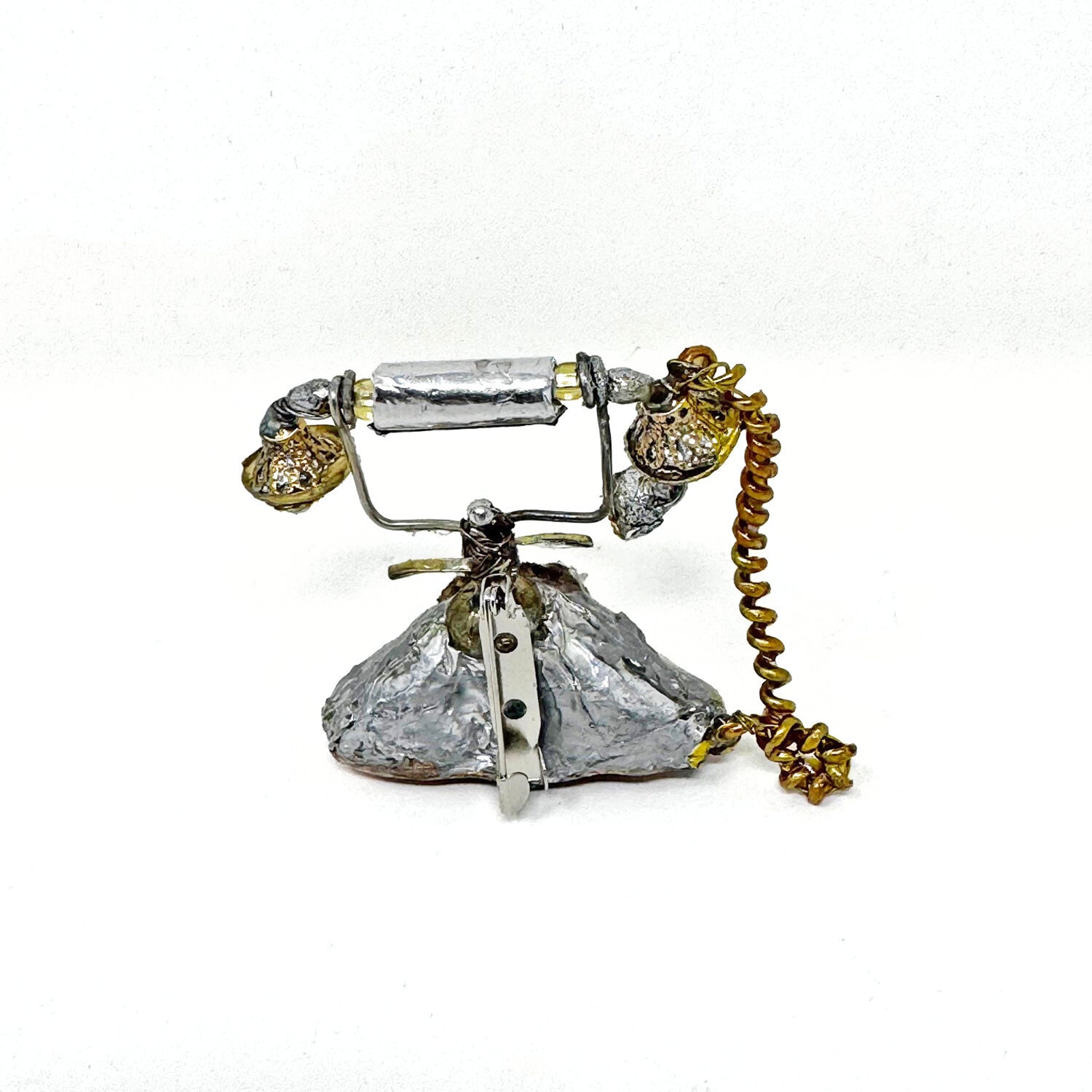 Backside of a mini sculpture of a silver antique style rotary phone, made of recycled materials against a white background. There is a metal pinback attached to the sculpture  that allows it to be worn as a brooch.