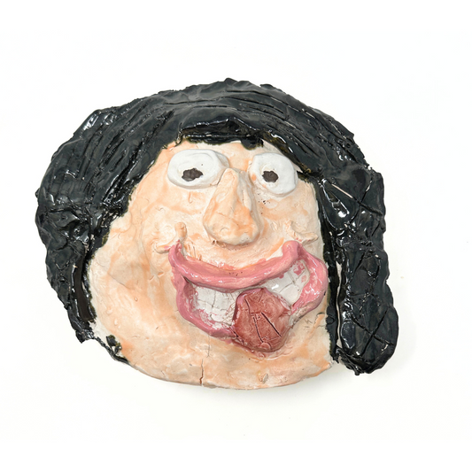 A ceramic sculpture of a head, flattened like a mask. The face has peach skin, 2 round white eyes, a triangular nose, and a grinning mouth with white teeth and a red tongue sticking out. The face is framed with dark black hair. 