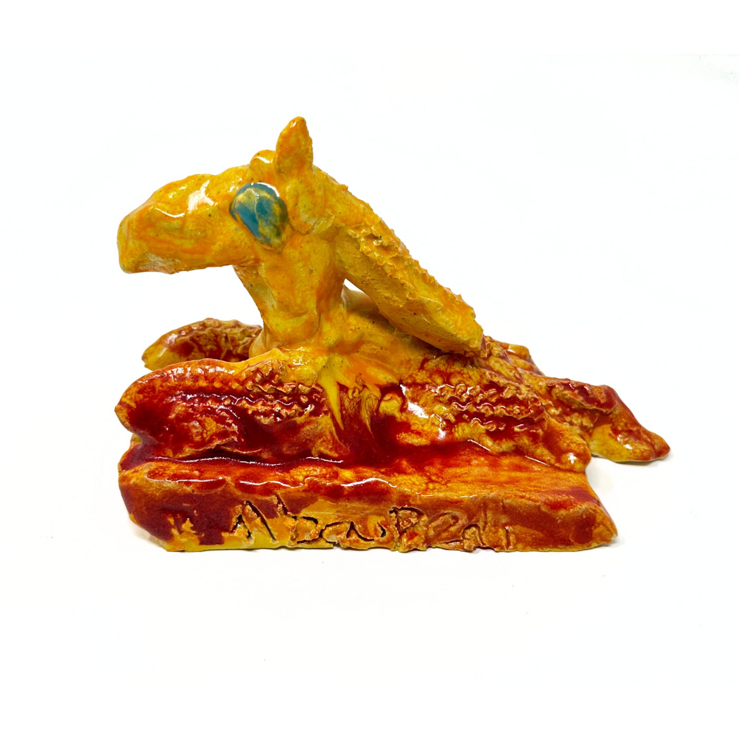 A small ceramic figurine modeled after a pokemon character. The creature is seated on a red square base, with the name "Bulbapedi" etched onto the side. The creature is glazed orange and red with a round light blue eye. It is shaped roughly like a seated horse or dragon.