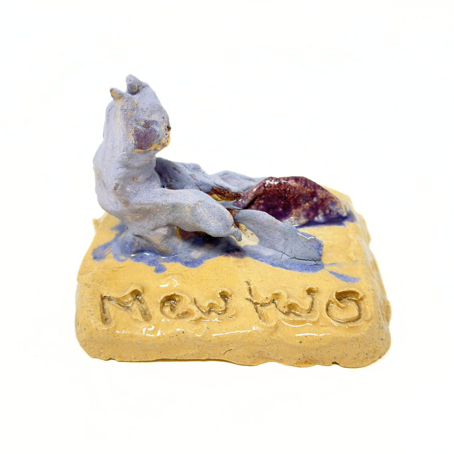 A glazed ceramic figurine modeled after a pokemon character. The figure is shaped like a cat, glazed a matte purple. It sits on a yellow square base which has the name "Mew Two" etched into its surface. 