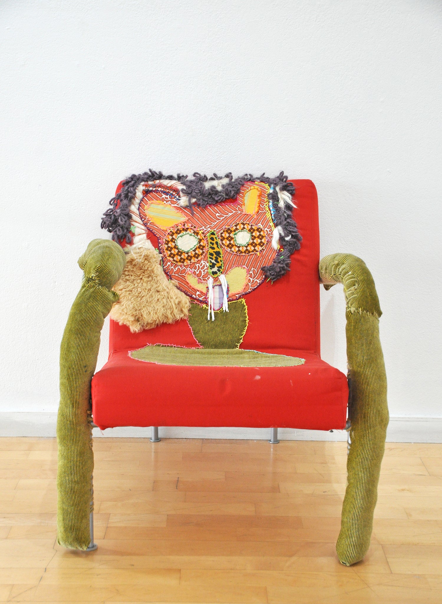 a sculpture of a lion chair made using a found red chair and various textiles. The artist has sewn a lion face onto the chair back and covered the arms in soft brown corduroy fabric.