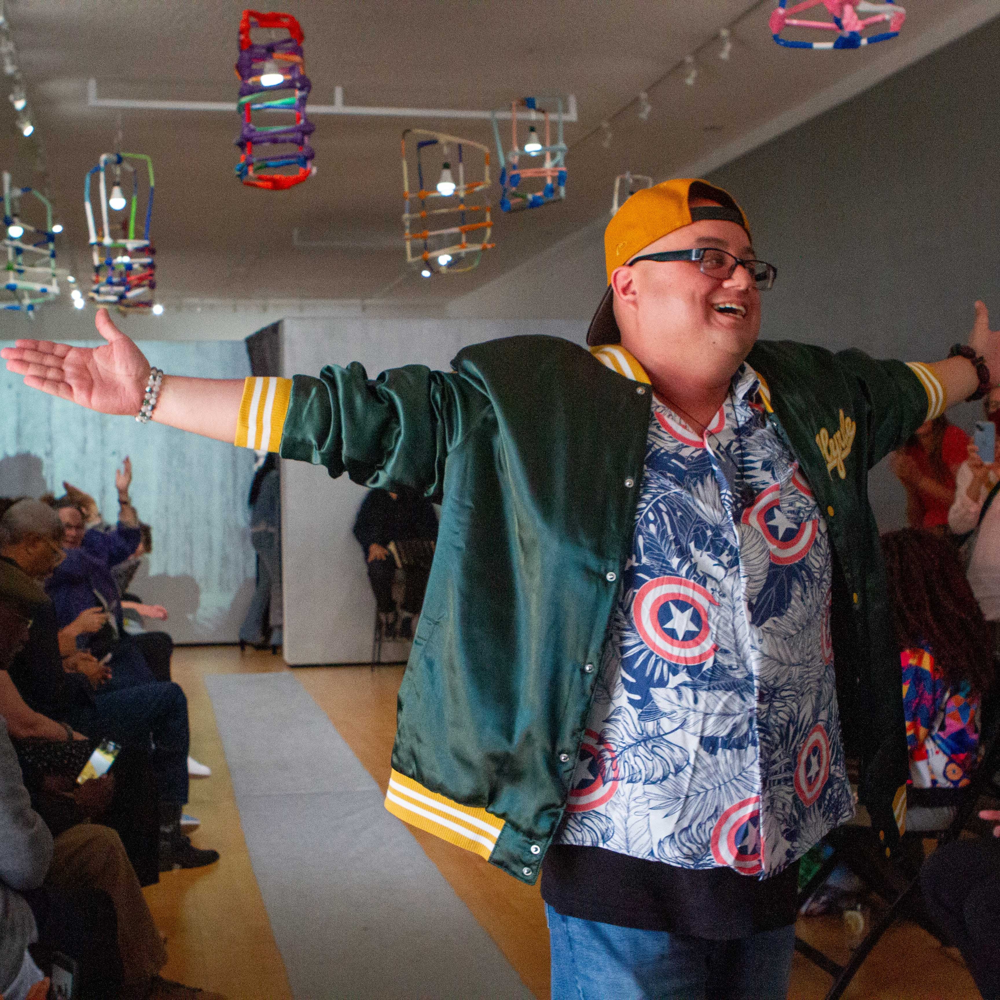 A photograph of a model walking down a silver fashion runway. The model is artist Saul Alegria, a person with light skin who wears glasses, a backwards yellow cap, a green jacket, and a patterned blue and red shirt. Saul extends both arms in a confident gesture. The crowd seated behind Saul is smiling, applauding, and taking photographs. The walls are painted silver and there are colorful paper lanterns hanging from the ceiling. 