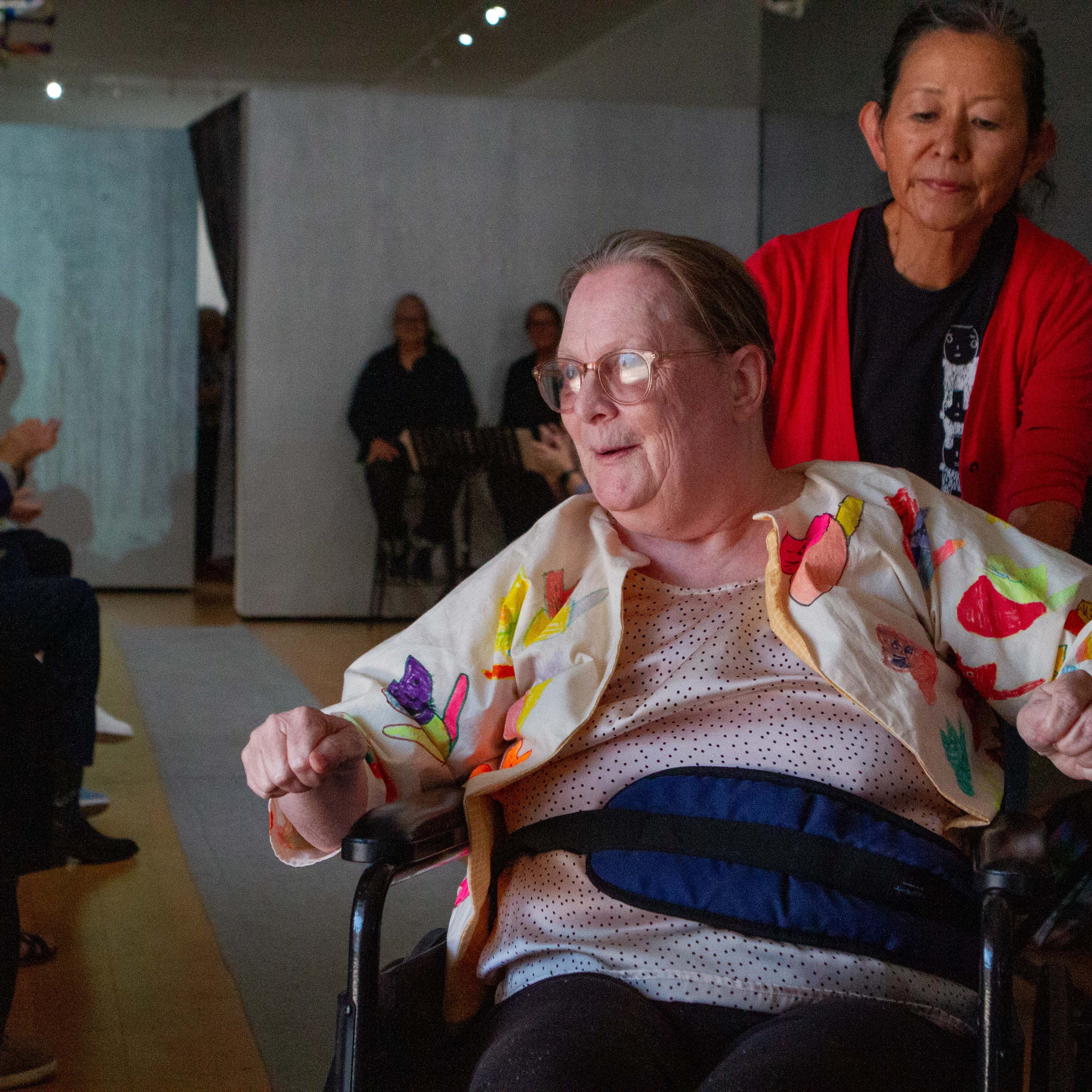 A photograph of a model in a wheelchair on a fashion runway. The model is artist Ann Meade, a person with white skin, straight light brown hair, who is wearing a white jacket with colorful hand-drawn cat designs and a black and white polka-dot shirt. A person wearing a red cardigan pushes Ann's wheelchair. The crowd on either side of the runway is clapping and smiling. The walls are painted silver. 