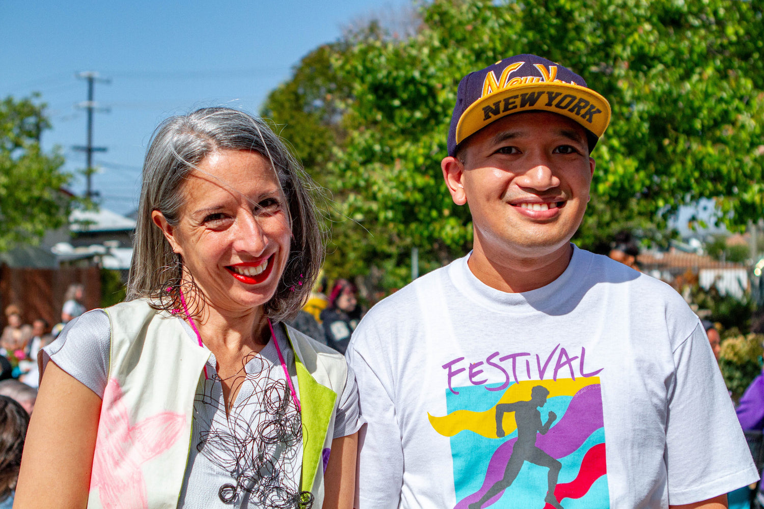 An image of two people standing outside on a sunny day and smiling at the camera. On the left is NIAD's executive director, Amanda Eicher, a woman with white skin and straight grey hair, wearing a statement wire necklace and vest with hand drawn designs. On the right is artist Danny Thach, a person with light skin and close cropped hair, wearing a purple and yellow baseball cap and a teeshirt with a colorful design. There are green trees and a crowd of people in the background. 