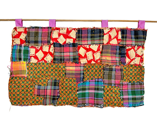 A long rectangular quilt made up of different square fabric swatches, hanging on a wooden dowel from 4 purple fabric loops. The patterns of the fabric swatches are purple plaid, checkered green and orange fabric, and a red fabric with white square designs. The quilt is hand sewn with a running stitch in yellow and purple thread. 