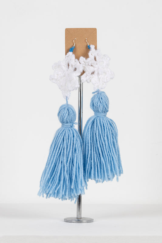 A pair of crocheted earrings hanging from on a tan card, photographed against a white background. The earrings are white yarn snowflakes with dramatic large blue tassels. They have thin silver hooks.
