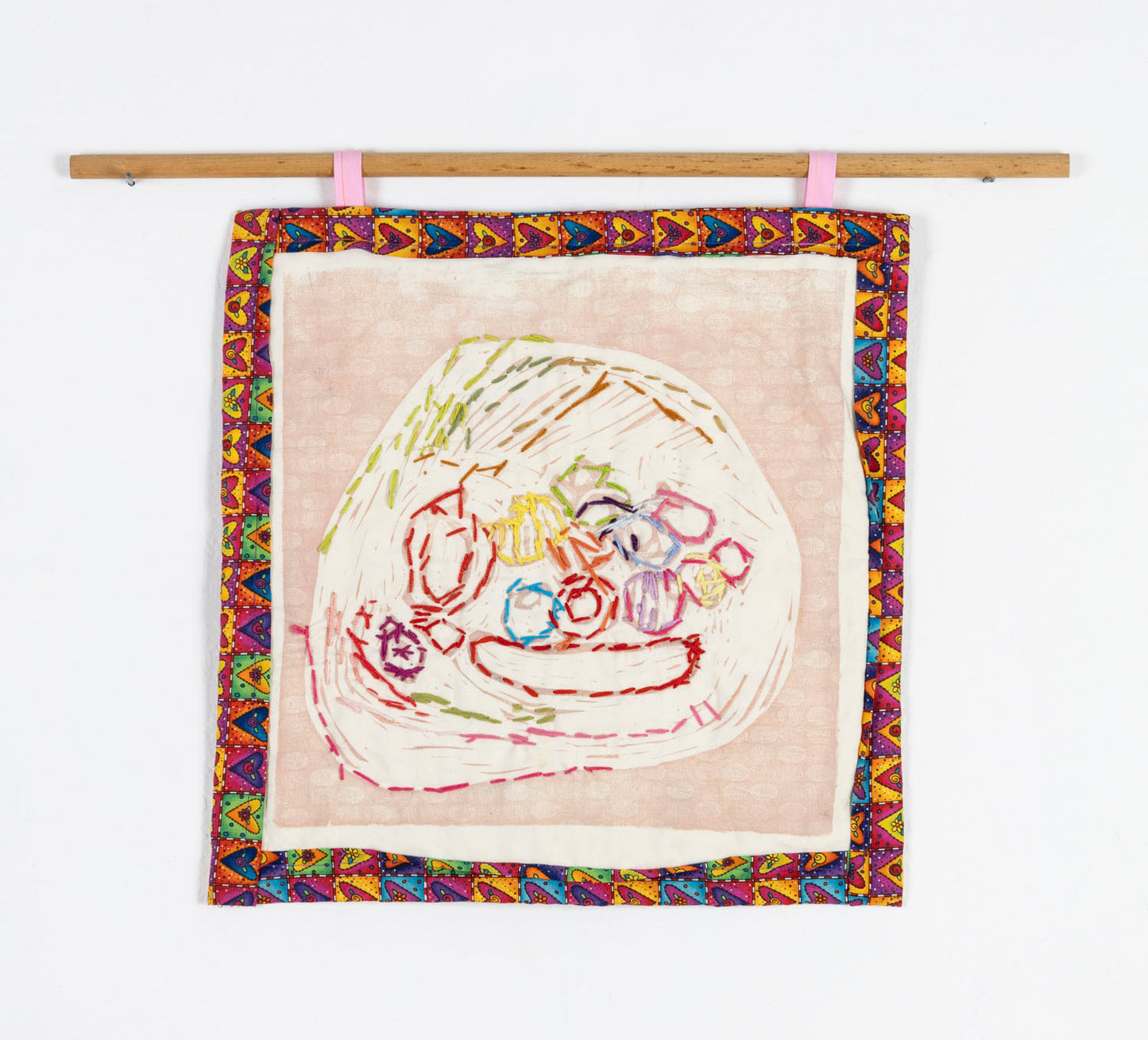 An embroidered print on fabric, hanging from a wooden dowel. Using pale pink ink, the artist has printed a still life on tan fabric. The objects in the still life, various fruit, have been embroidered over with rainbow colored thread. The print is bordered with a band of heart patterned fabric.