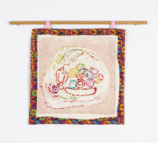 An embroidered print on fabric, hanging from a wooden dowel. Using pale pink ink, the artist has printed a still life on tan fabric. The objects in the still life, various fruit, have been embroidered over with rainbow colored thread. The print is bordered with a band of heart patterned fabric.