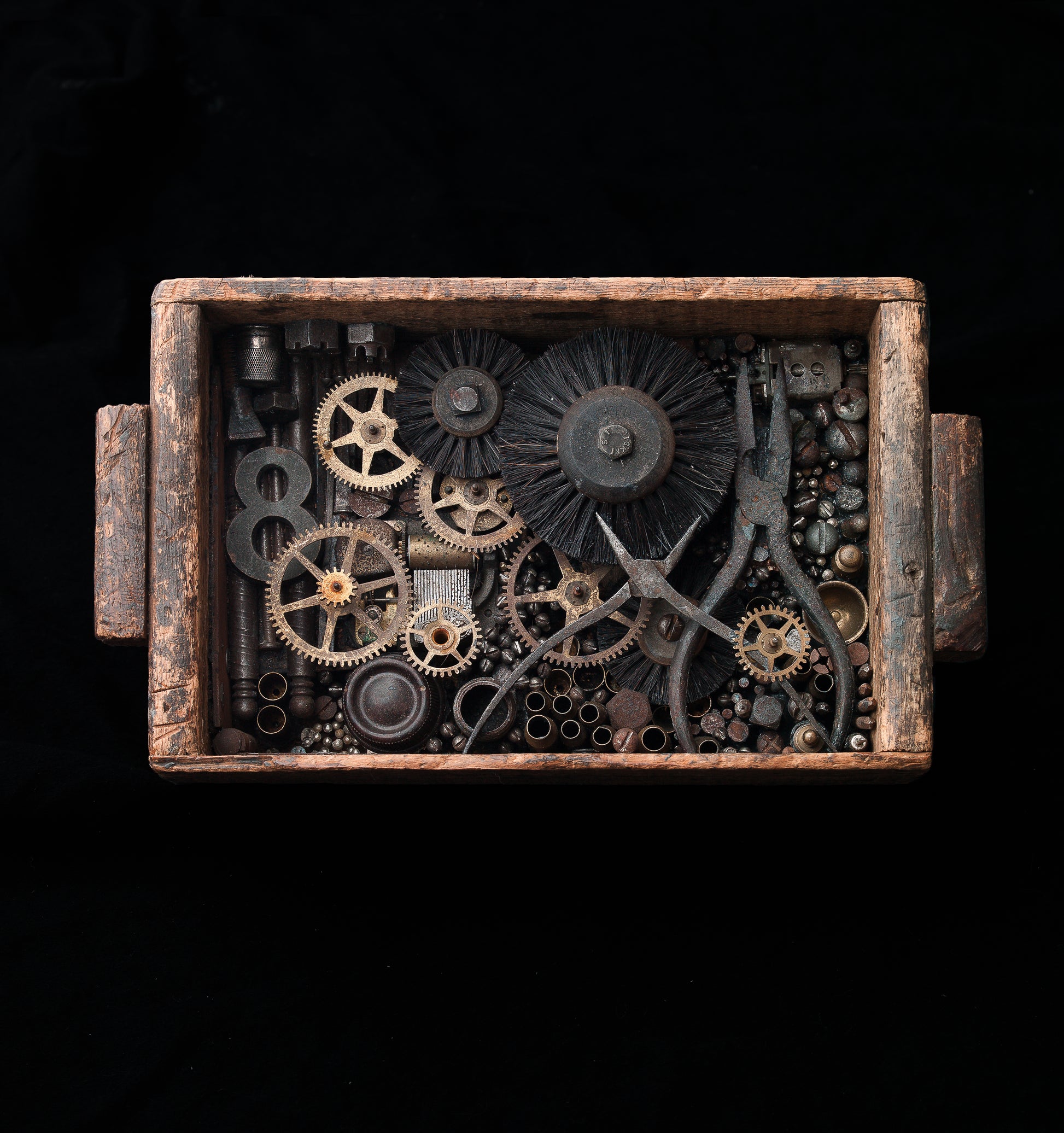 Assemblage made out of found materials in a rustic wooden box. The items in the box include gears, pliers, sanding wheels and brushes, and other aged metal ephemera. The box has a wire on the back for hanging. 
