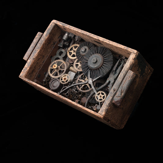 Assemblage made out of found materials in a rustic wooden box. The items in the box include gears, pliers, sanding wheels and brushes, and other aged metal ephemera. The box has a wire on the back for hanging. 