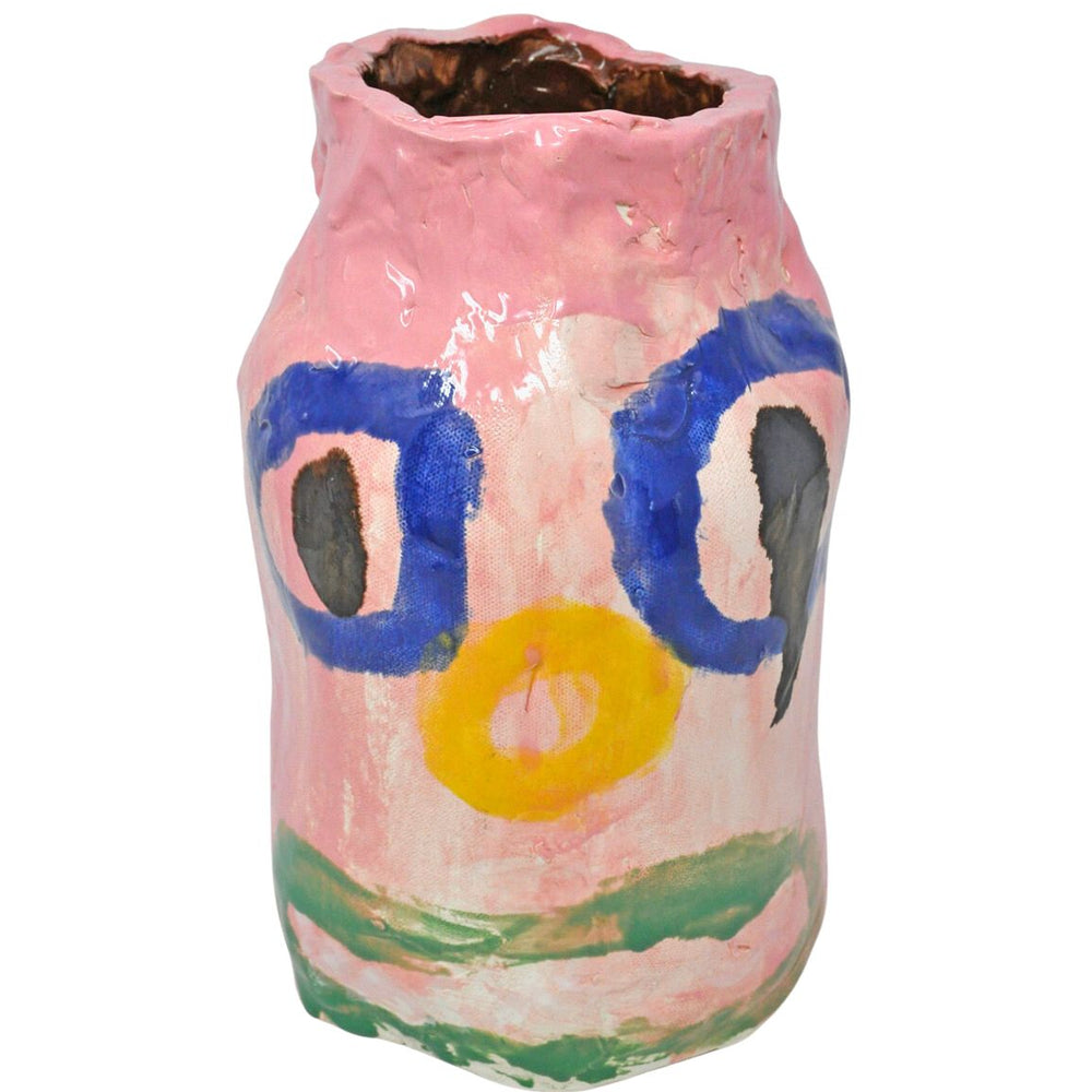 A ceramic vessel photographed in a white space. The vessel is a tall pink vase with a clown face painted on the front. The face has large blue circle eyes, a yellow circle nose, and a wide green mouth. The interior of the vase is glazed dark brown.