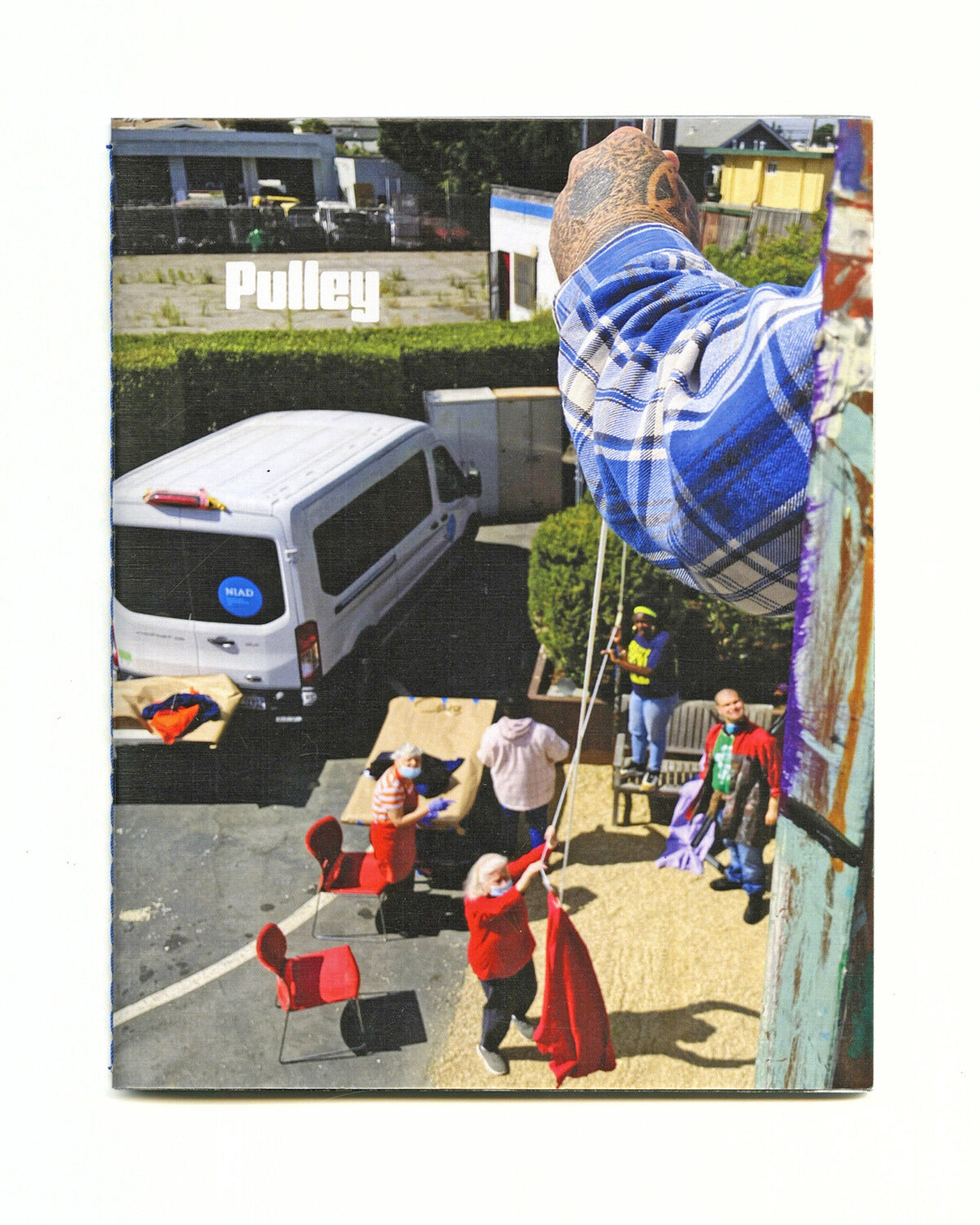 Pulley exhibition catalog cover, which features a tattooed hand lowering fabric downwards on a pulley. Artists and the NIAD van can be seen below. 