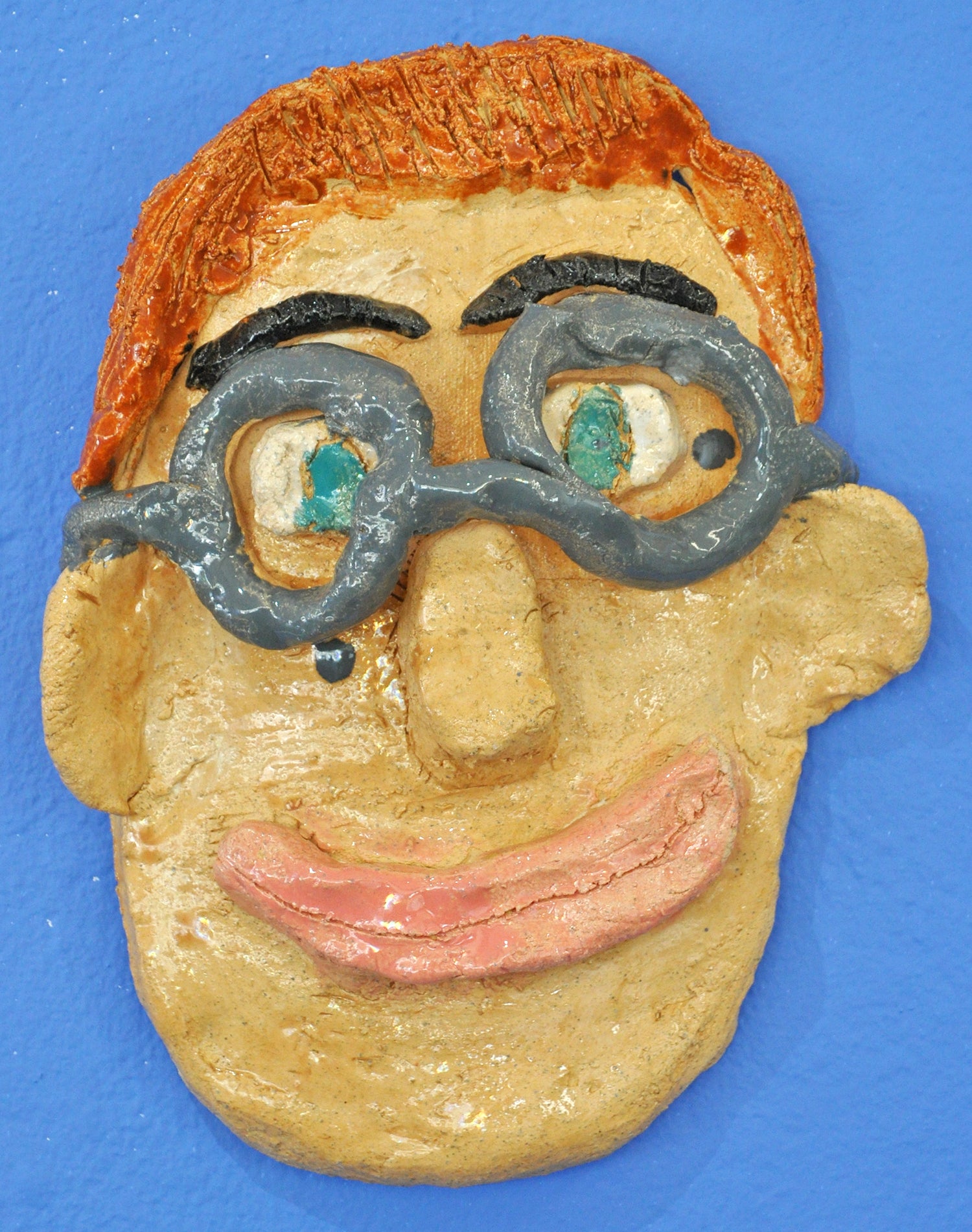 A face made of ceramic, against a blue wall. The face has green eyes, medium toned skin, red har, and large-rimmed gray glasses. Above the glasses are black eyebrows. The face is smiling. 