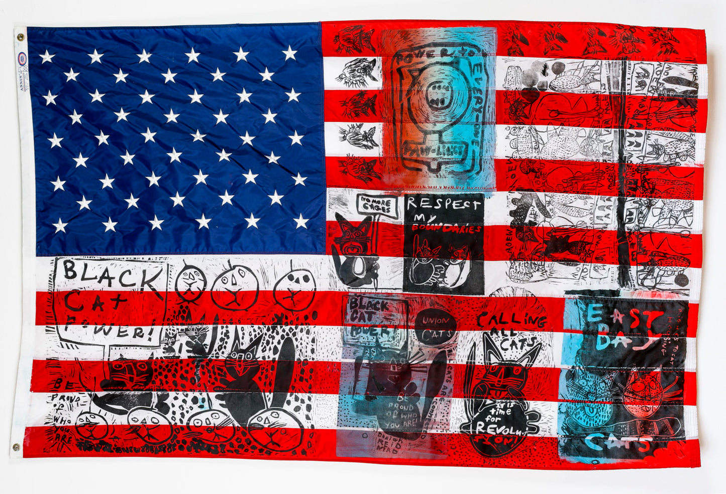 A large fabric American flag with black ink linocut prints stamped across its surface. The printed imagery features cats, pumpkins, symbols, and empowering phrases like: Black Cat Power, Union Cats, Respect My Boundaries, and Power to Everyone. 