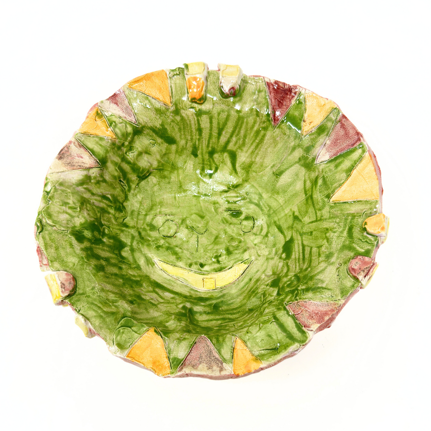 a ceramic bowl, photographed from above so that the interior surface is in full view. The interior is glazed all bright green, with yellow, red, and orange triangles sculptured around the outer rim. At the base of the bowl, the artist has etched a smiling face with a wide open mouth that has a single gold tooth. 