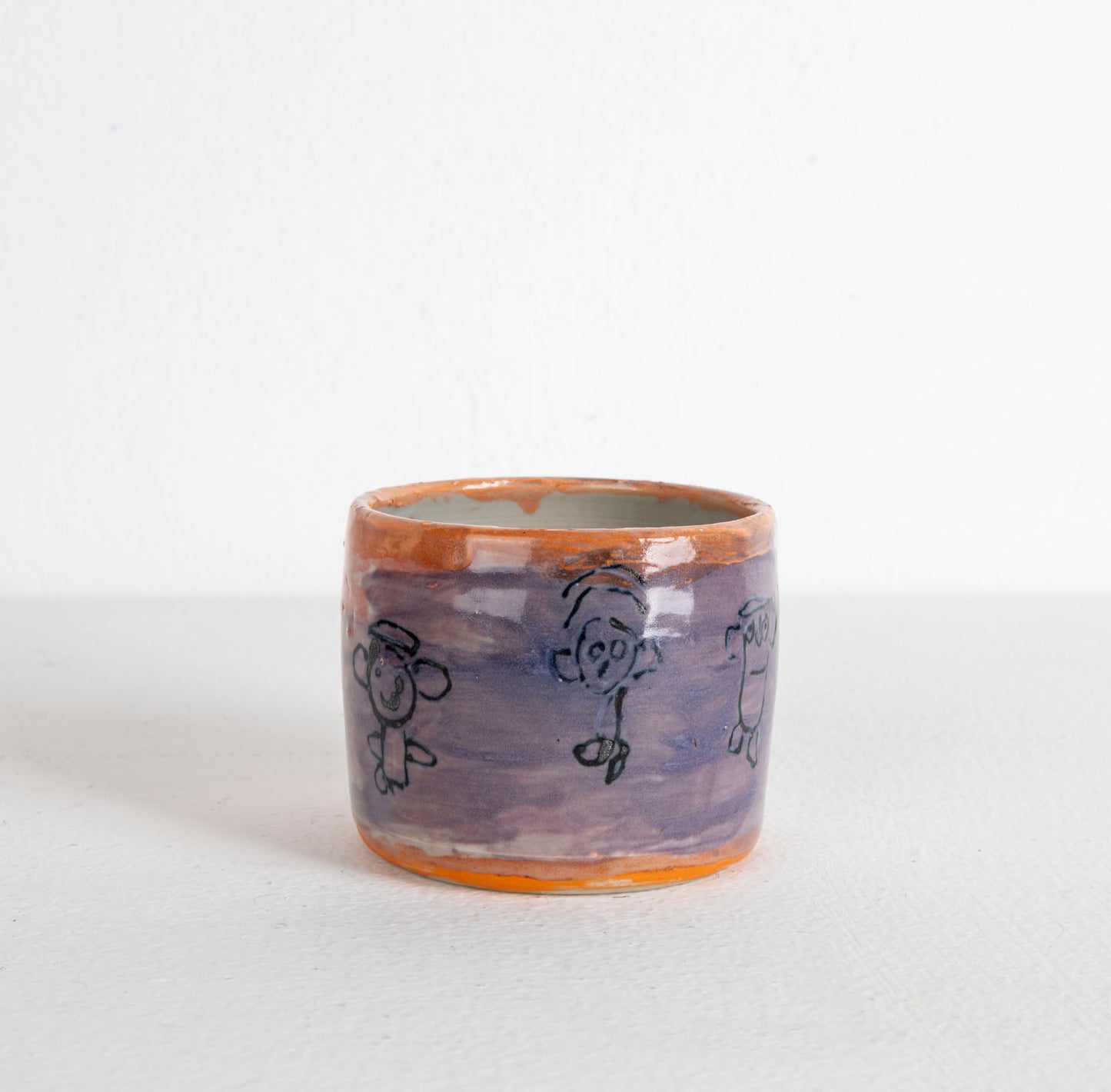 A small wheel-thrown ceramic cup, glazed with glossy purple, white, and orange. There are little figures painted around the outer surface. 