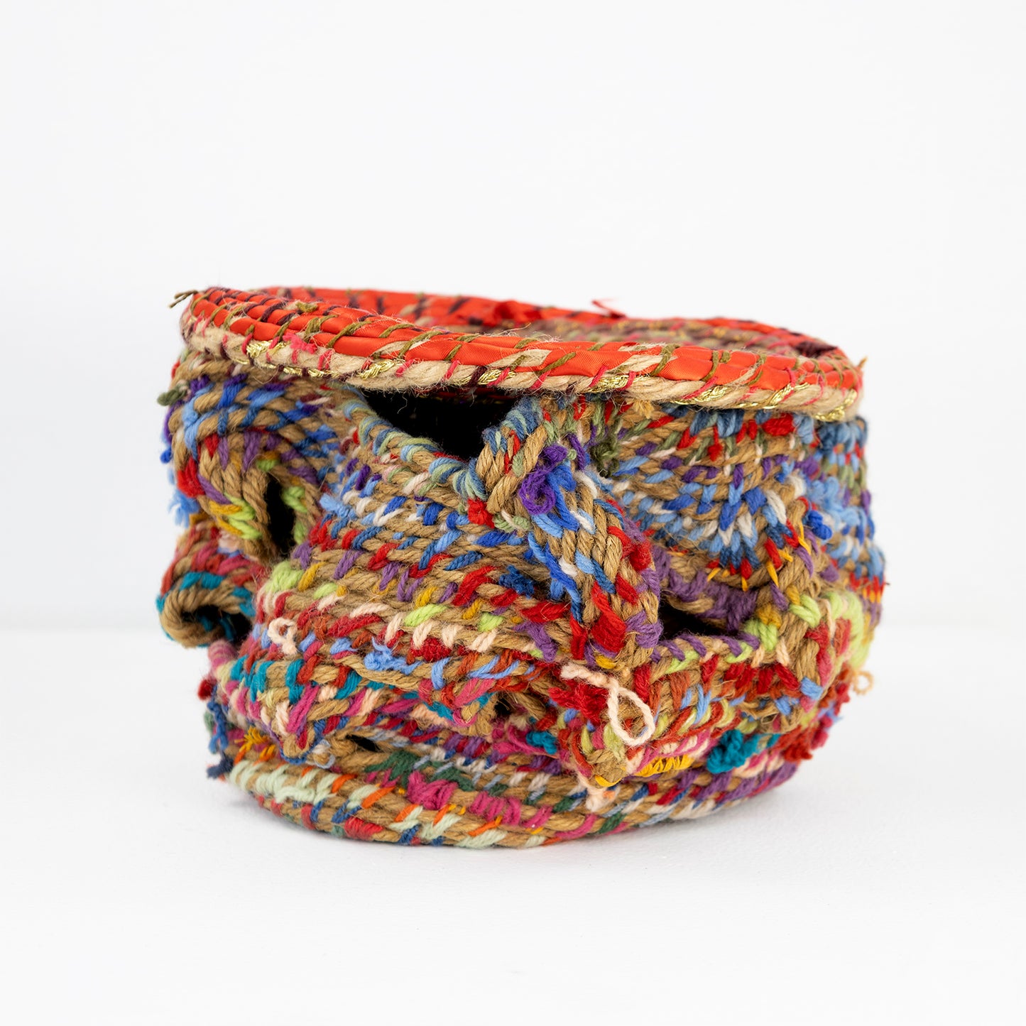 A colorful handcrafted basket with an orange lid. The basket is intricately woven with thick, multicolored yarn, featuring vibrant hues of red, blue, yellow, green, and purple. The weaving creates a textured, layered appearance with visible gaps and overlaps in its form.