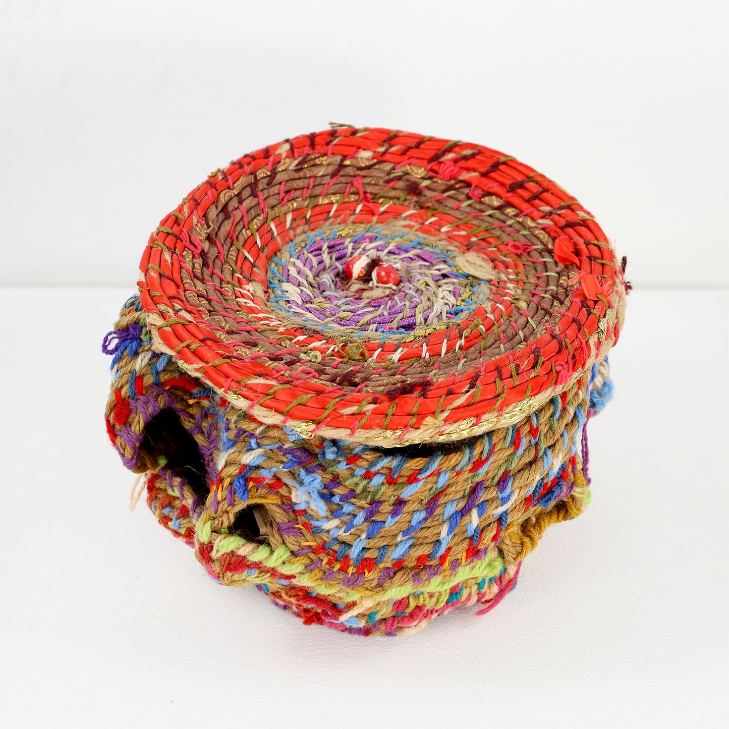 A colorful handcrafted basket with an orange lid. The basket is intricately woven with thick, multicolored yarn, featuring vibrant hues of red, blue, yellow, green, and purple. The weaving creates a textured, layered appearance with visible gaps and overlaps in its form.  The basket's lid and body are tightly woven with a variety of colorful fibers, including red, purple, brown, blue, and green. The top of the lid features a circular pattern with a central point, predominantly red with accents of purple.
