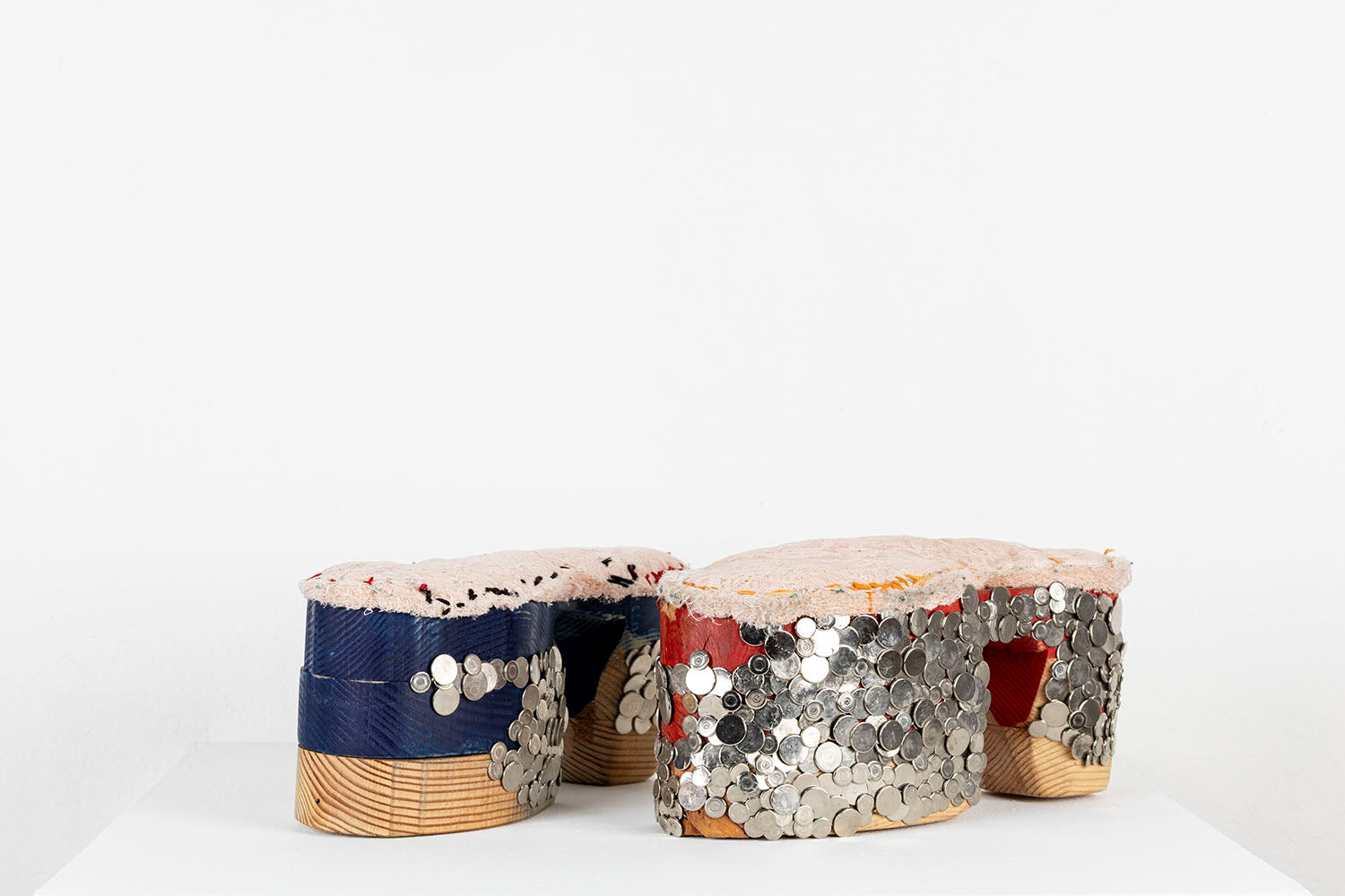 A mixed media sculpture resembling a pair of platform shoes. The base of the shoes are stacked wooden blocks that have been carved and rounded over. One shoe has been partially painted blue and the other has been partially painted red. The artist has hammered dozens of flat, shiny metal tacks into the wood outer sole, in a pattern that resembles chainmail armor, grommets, or fish scales. Lacey pink fabric has been stitched along the top of each shoe with blue, red, or orange thread. 