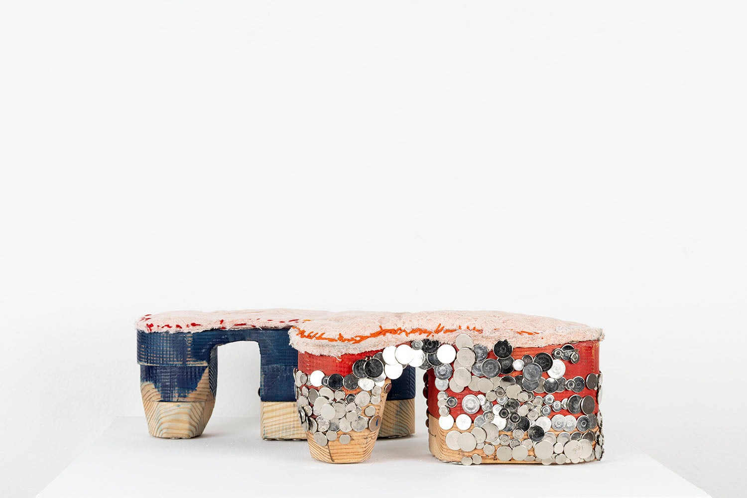 A mixed media sculpture resembling a pair of platform shoes. The base of the shoes are stacked wooden blocks that have been carved and rounded over. One shoe has been partially painted blue and the other has been partially painted red. The artist has hammered dozens of flat, shiny metal tacks into the wood outer sole, in a pattern that resembles chainmail armor, grommets, or fish scales. Lacey pink fabric has been stitched along the top of each shoe with blue, red, or orange thread. 