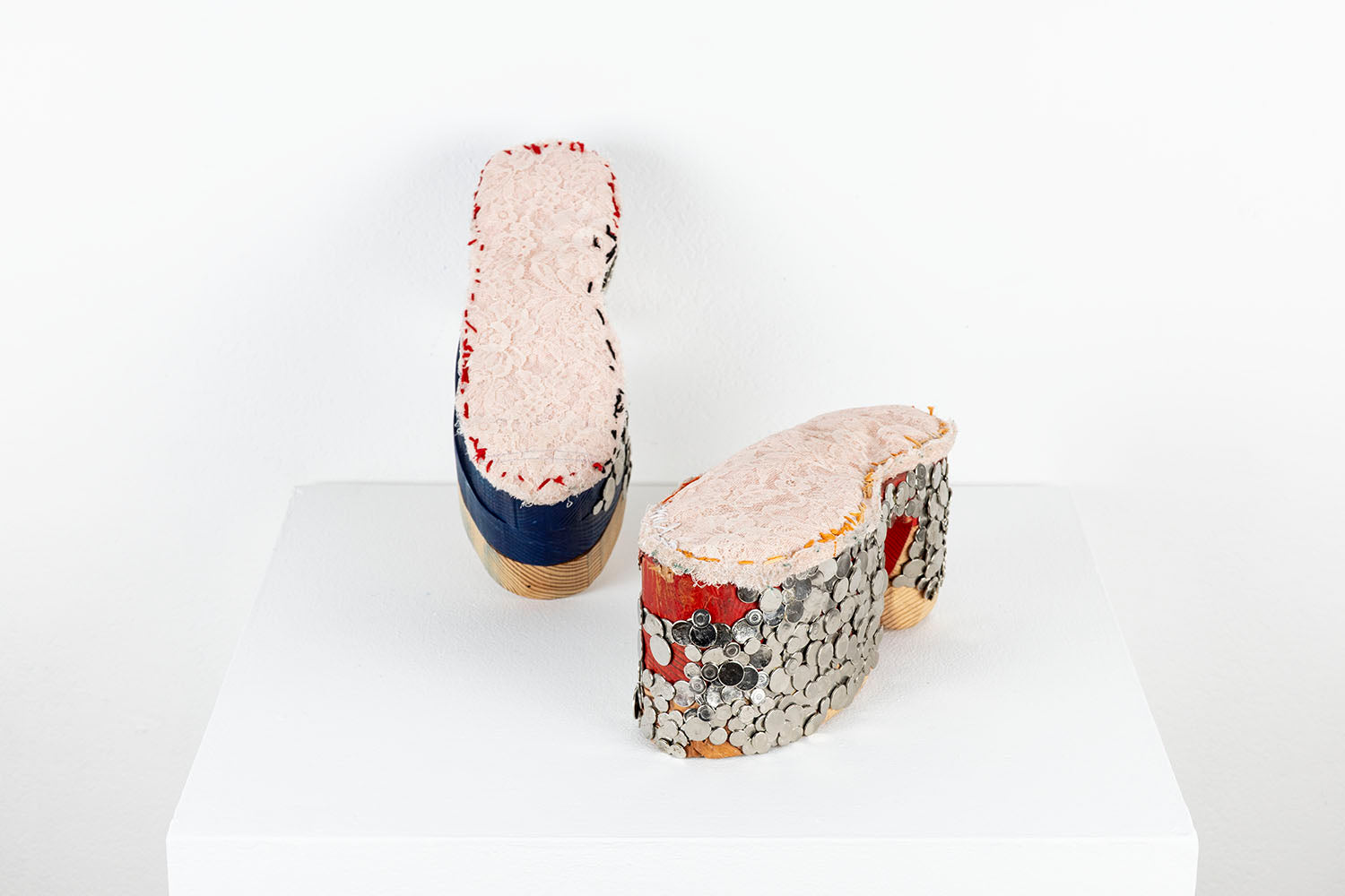 A mixed media sculpture resembling a pair of platform shoes. The base of the shoes are stacked wooden blocks that have been carved and rounded over. One shoe has been partially painted blue and the other has been partially painted red. The artist has hammered dozens of flat, shiny metal tacks into the wood outer sole, in a pattern that resembles chainmail armor, grommets, or fish scales. Lacey pink fabric has been stitched along the top of each shoe with blue, red, or orange thread. 
