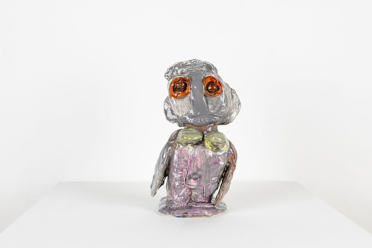 A small ceramic figurative sculpture, based on an African doll. The sculpture is glazed with shiny grey layered over violet and green. It has large round brown eyes which stand out in stark contrast with the muted tones on the rest of the body. It leans slightly toward the right. The figure is positioned on a flat ceramic base that is shaped like a face, as if the doll is emerging from the face's mouth. 