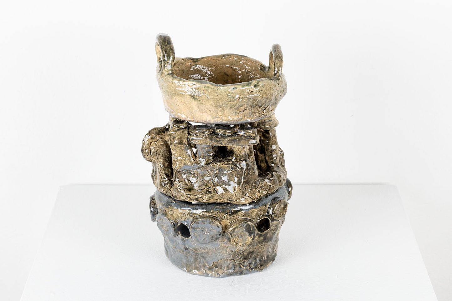 The same sculpture photographed from a slightly different angle. The form is three stacked objects that represent a handled pot on a traditional stove. The form on the bottom features a ring of round protrusions and holes. The form in the middle has intersecting vertical and horizontal cylinders. And the form on the top appears to be a deep pan with two looped handles. 