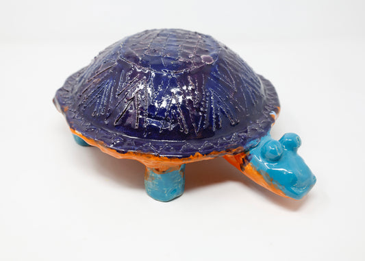 A ceramic turtle that has a dark purple-blue glazed shell embellished with engravings. The body of the turtle is a bright orange with red and blue glaze splatters. The head and legs are bright blue.