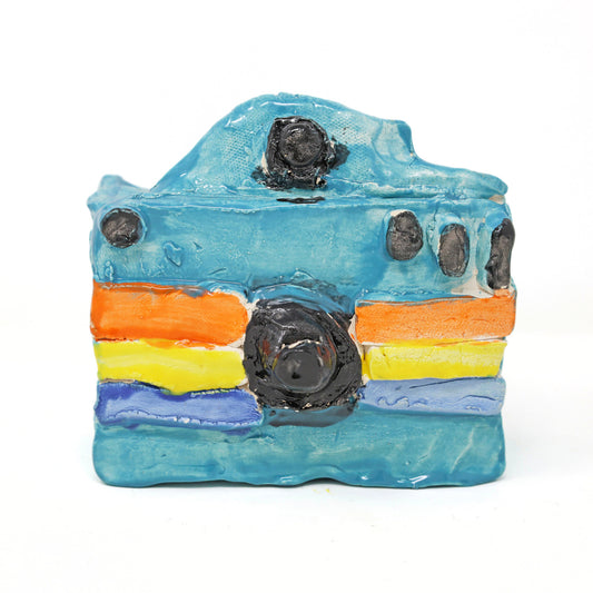 This is a ceramic sculpture of a camera. The camera body is glazed in a bright blue color, with the lens and buttons highlighted with a black glaze. There are three colored stripes that go horizontally across the camera, in orange, yellow and dark blue. 