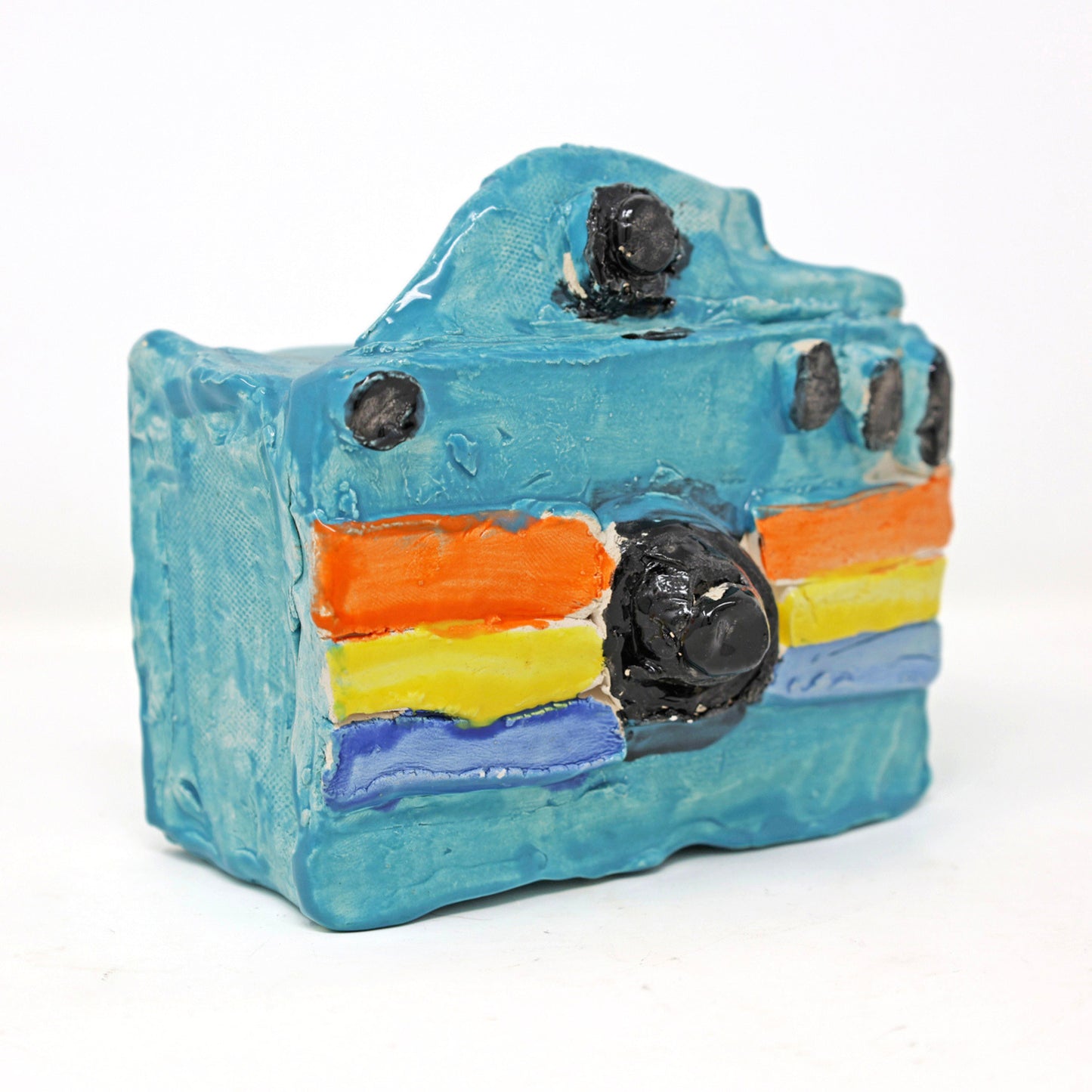 This is a ceramic sculpture of a camera. The camera body is glazed in a bright blue color, with the lens and buttons highlighted with a black glaze. There are three colored stripes that go horizontally across the camera, in orange, yellow and dark blue. 