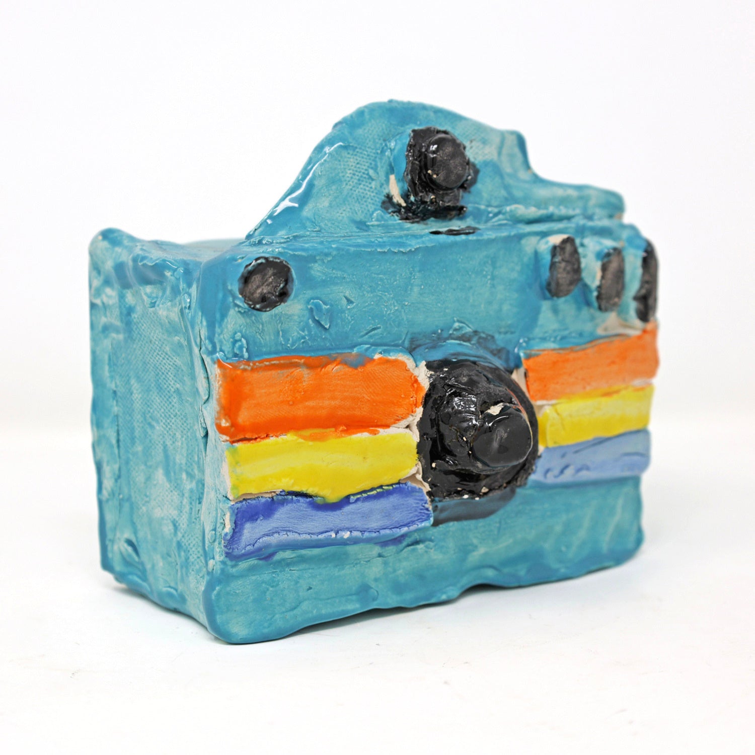 This is a ceramic sculpture of a camera. The camera body is glazed in a bright blue color, with the lens and buttons highlighted with a black glaze. There are three colored stripes that go horizontally across the camera, in orange, yellow and dark blue. 