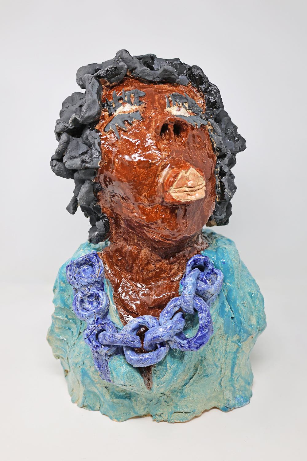 This is a ceramic sculpture of the top half of a figure. The figure is wearing a teal garment with a dark blue chained necklace. They have black curly hair and long, striking eyelashes. 