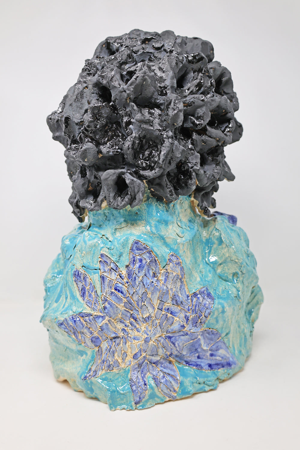 This is a ceramic sculpture of the top half of a figure. This image depicts the back of the figure, they have black curly hair and a dark blue flower on their teal garment.