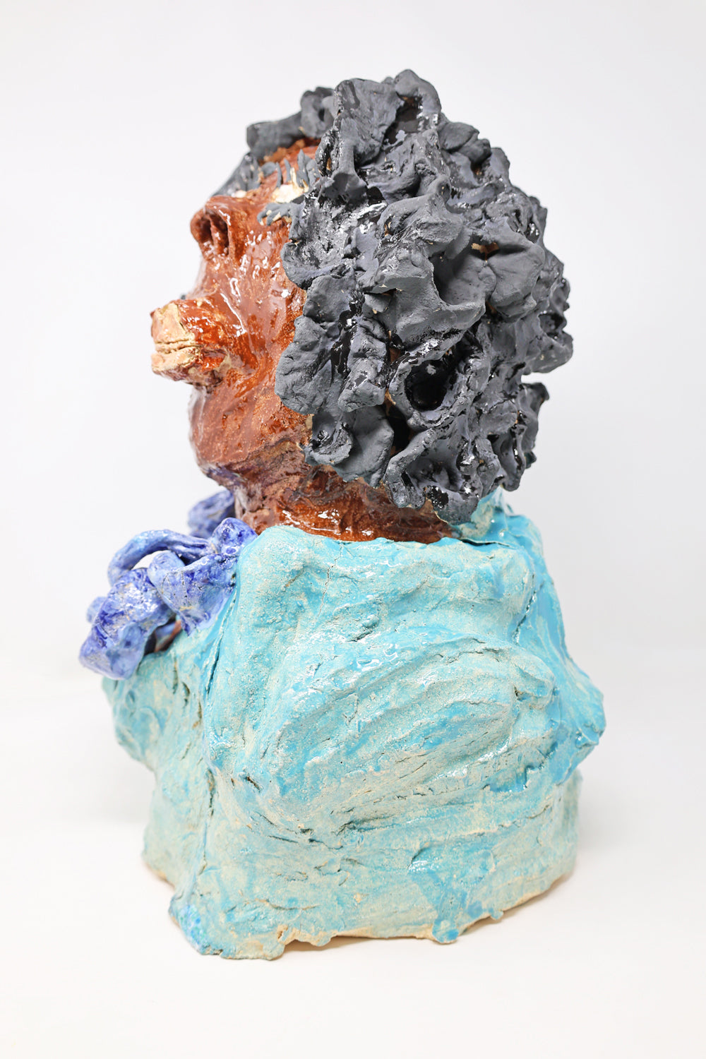 This is a ceramic sculpture of the top half of a figure. This image depicts the side profile of the figure. They have black curly hair, are wearing a teal garment with a chunky blue necklace.