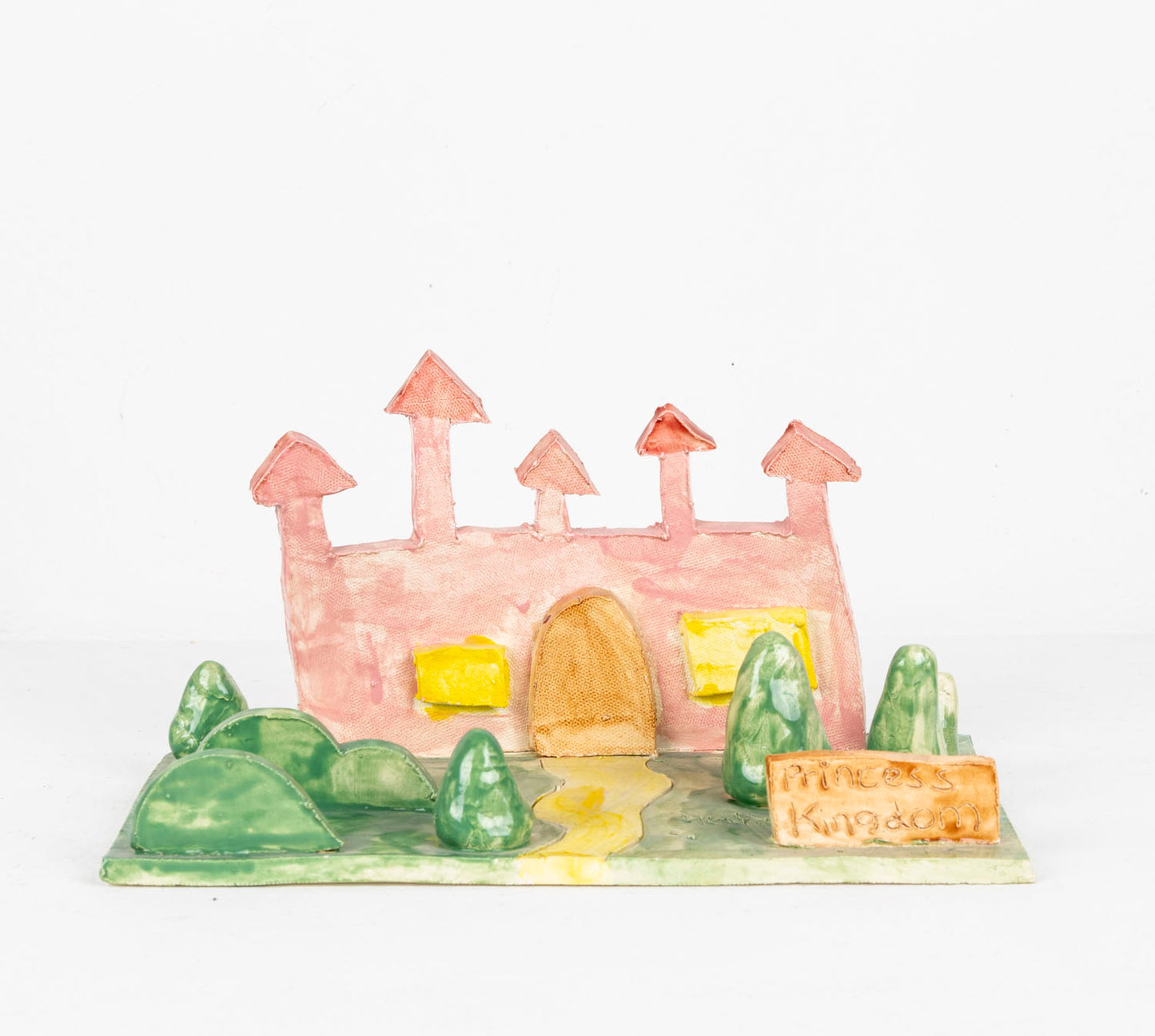 A small ceramic diorama made from thin slabs of clay. A pink castle with two big yellow windows and a grand rounded door sits on a green field which sprouts little conical trees and bushes. A sign positioned at the end of a winding yellow path reads: Princess Kingdom. 