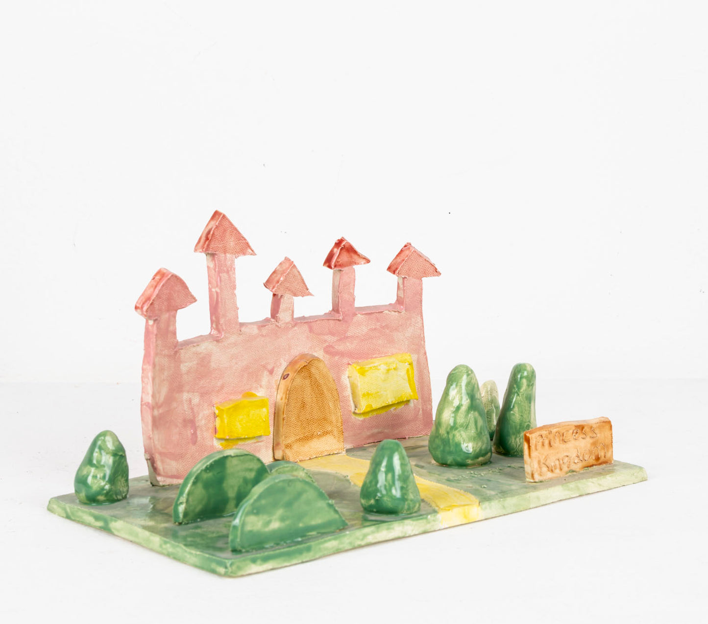 A small ceramic diorama made from thin slabs of clay. A pink castle with two big yellow windows and a grand rounded door sits on a green field which sprouts little conical trees and bushes. A sign positioned at the end of a winding yellow path reads: Princess Kingdom. 