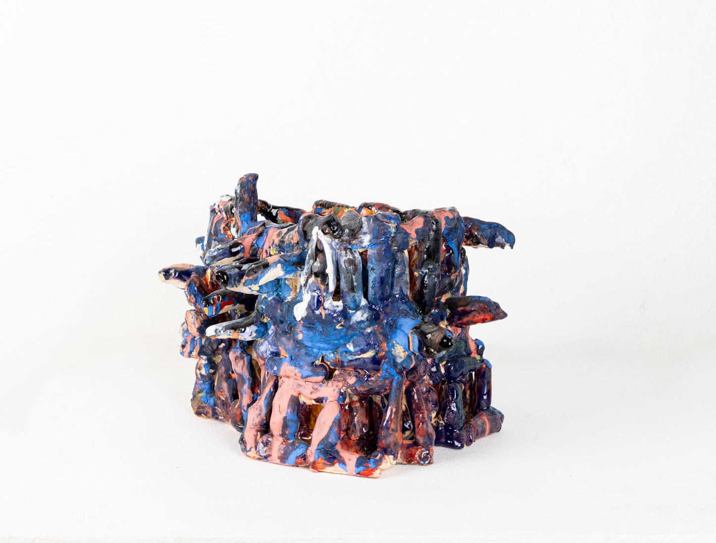 a ceramic sculpture of a two-tiered, cage-like structure glazed in shiny blue and matte pink. The structure is made of little cylinders, some are stacked vertically, others are wrapped around the form in horizontal bands, and several radiate out around the perimeter. There are gaps between each cylinder through which the white background is visible.